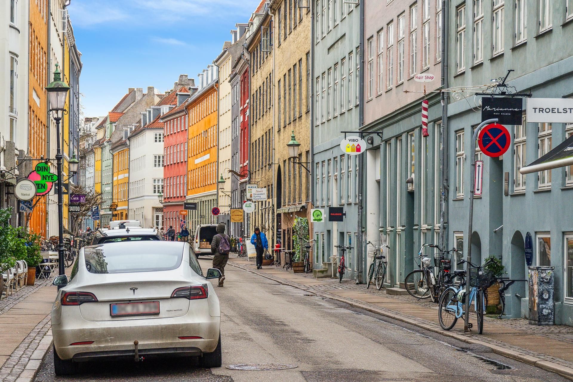 En Tesla Model 3 kører langs Sankt Peders Stræde, hvor farverige bygninger og cykler langs fortovet.