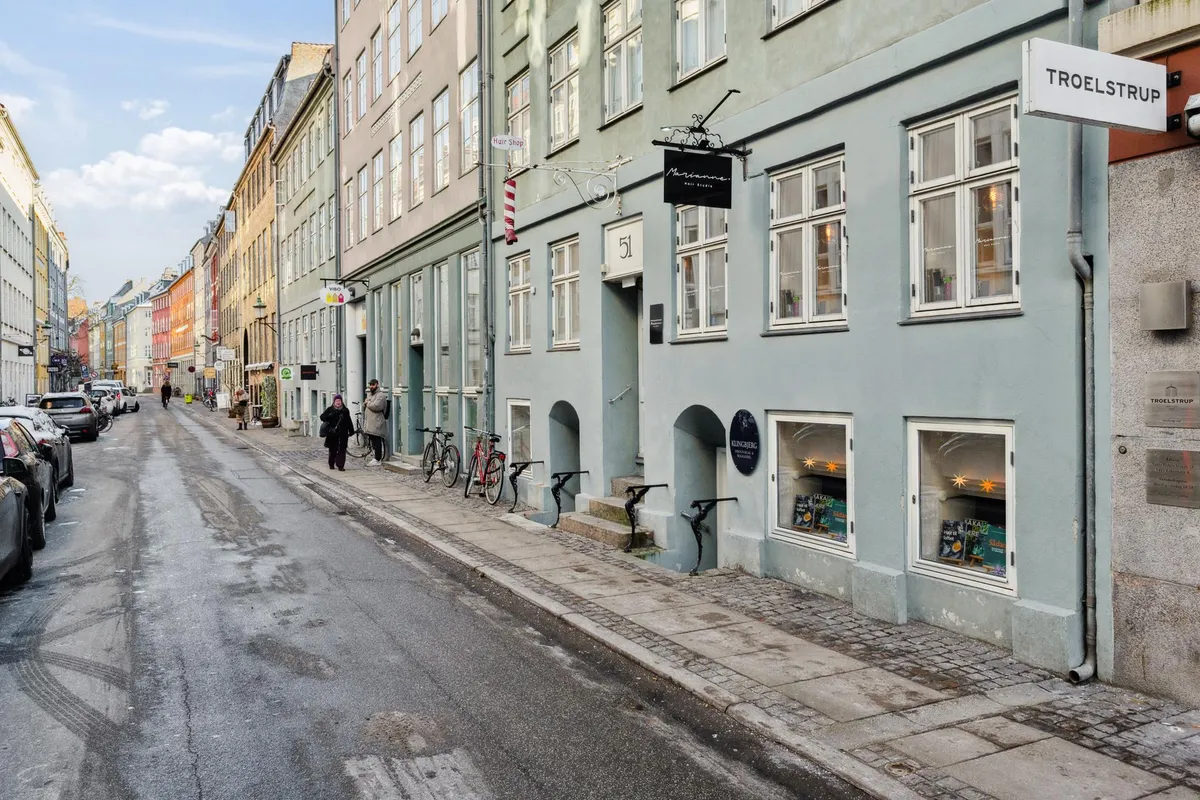 Gade med flere biler parkeret langs siderne, to butikker med vinduer og schilde, sammen med to personer og flere cykler