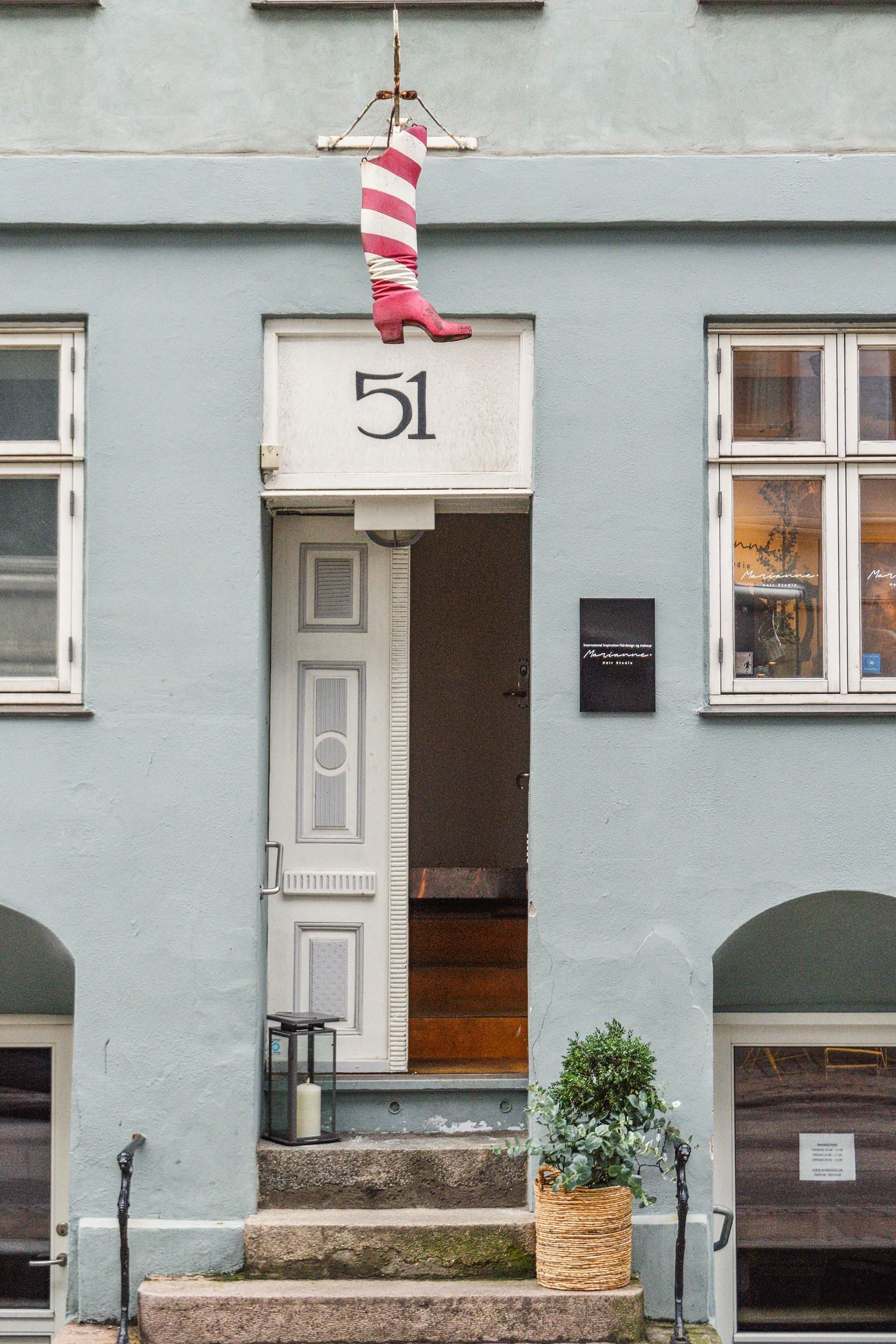 Lejlighedsdør med hængende rødt støvle og nummer 51, hvidt dørkarm og to vinduer, plante og en lysestage på trinene foran