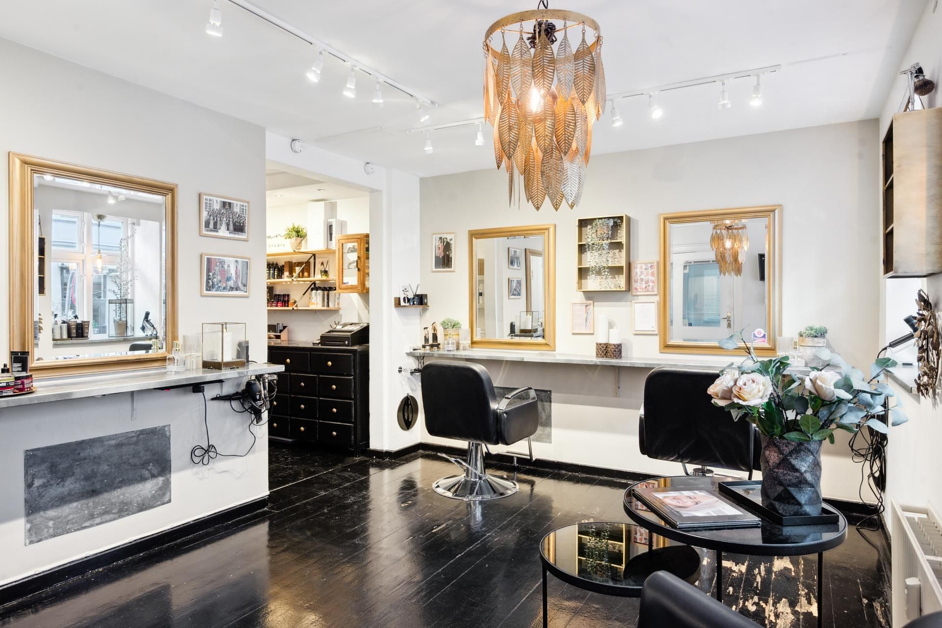 Indvendørs frisørsalon med lysegulle vægge og mørke trægulve, to stolpelyse og en lysekron, spejle og plante i potte og et