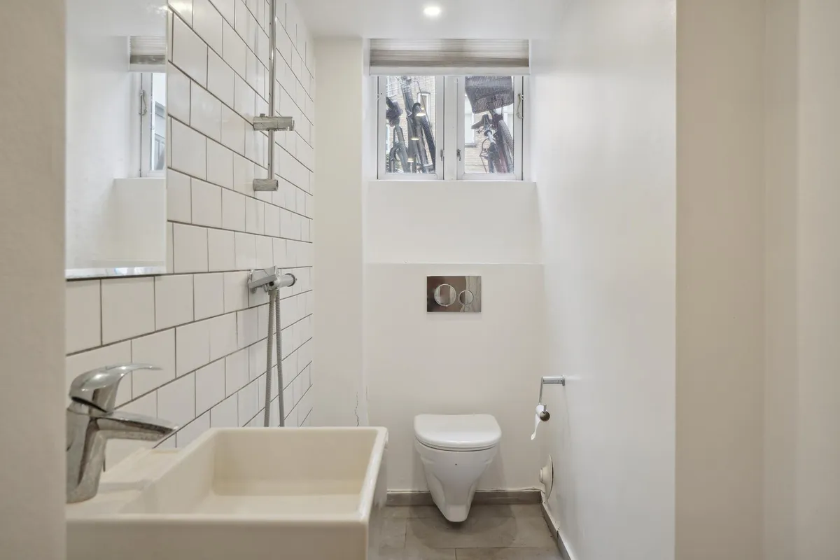 En hvid badeværelse med toilet, håndvask og vindue. Toilettet og håndvasken er moderne og hvidt. Væggene og gulvet er lavet