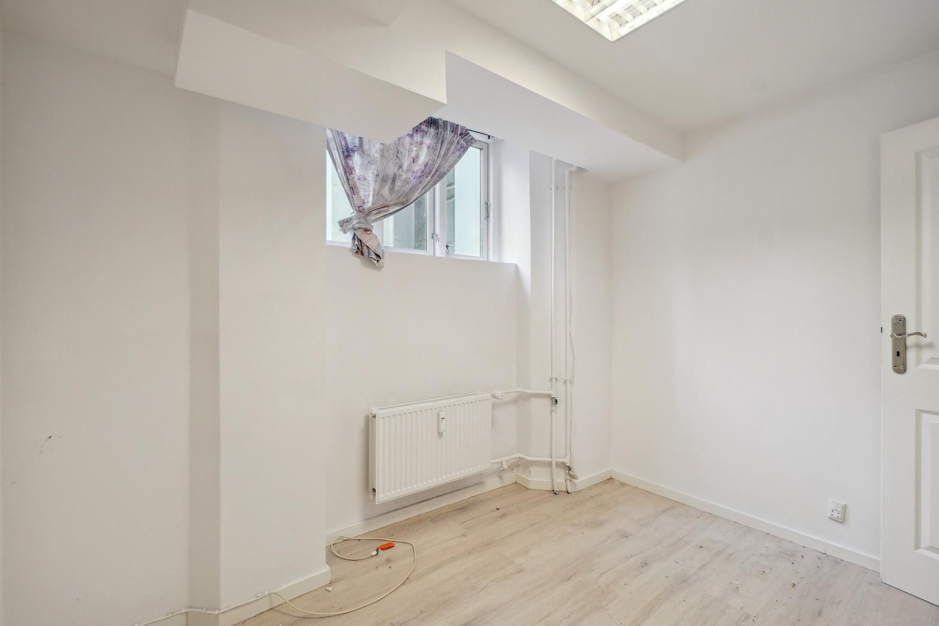 Kælderlejemål med moderne interiør, hvidt vægværk og trægulv samt radiator og enkle elektriske ledninger