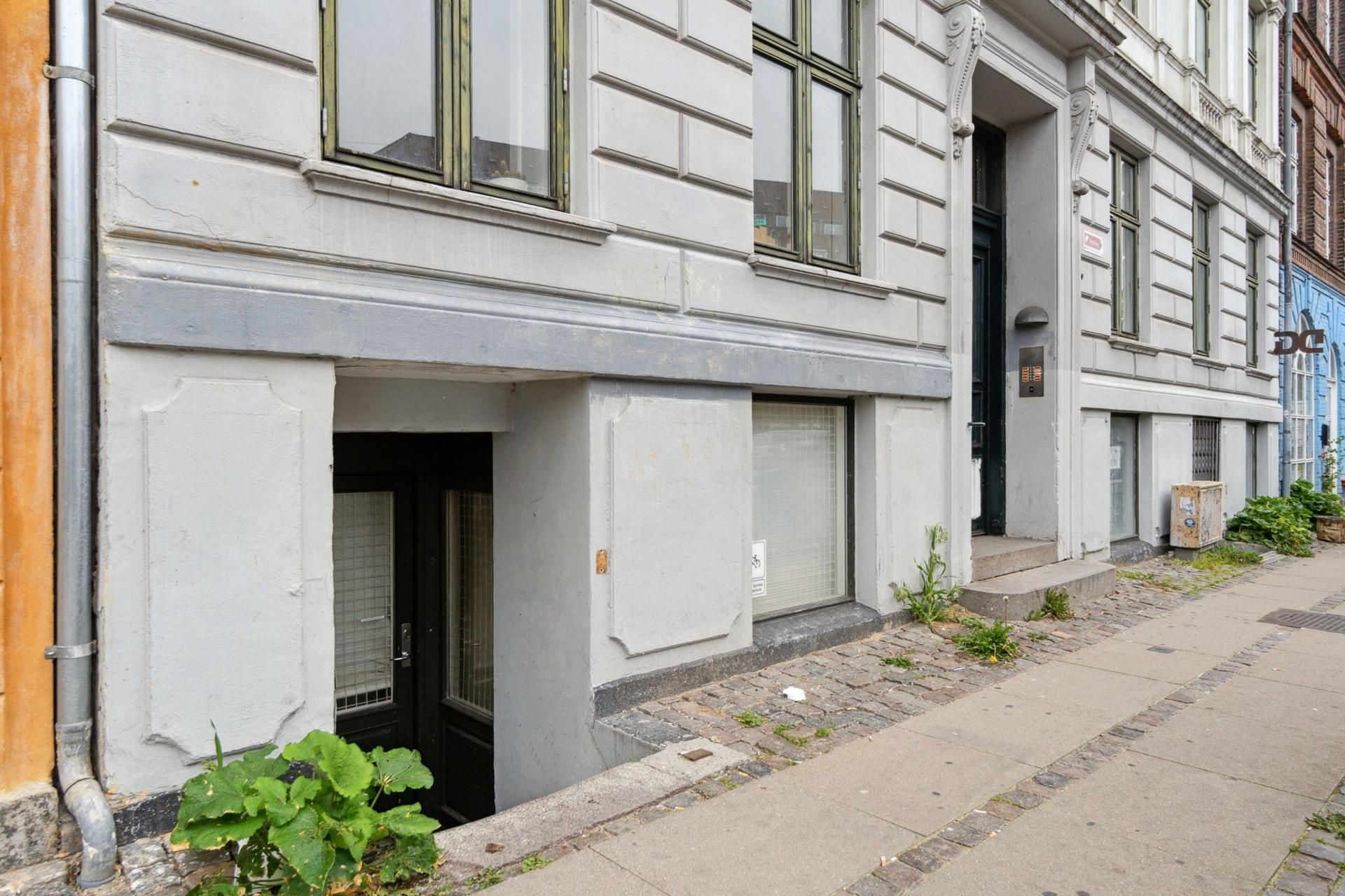 Kælderlejlighed med indgang til en smal gang og plantepotte til højre, foran en bygning med flere vinduer og døre.