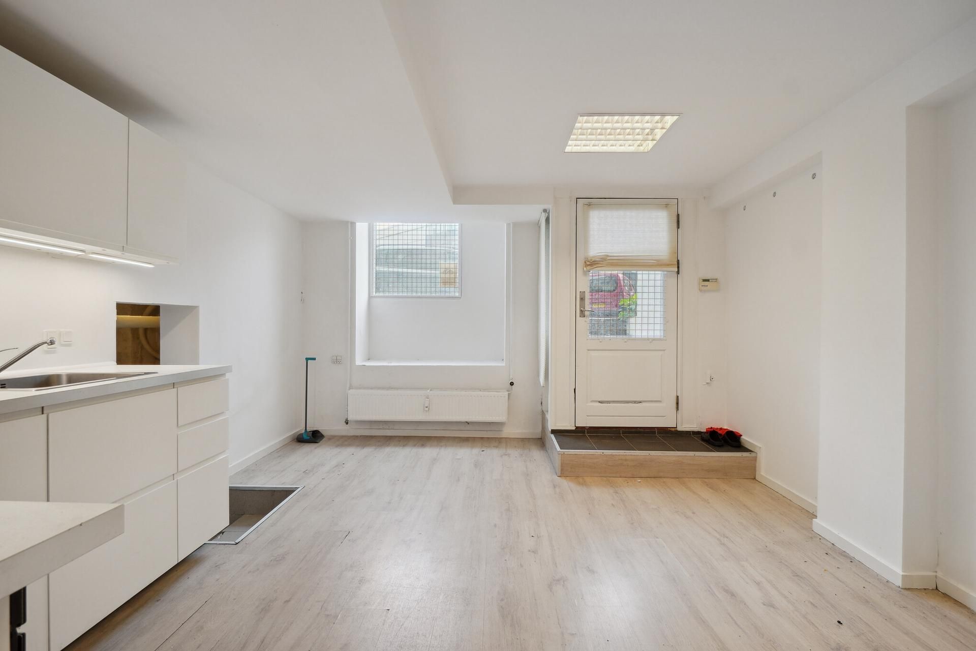 Lejlighed med lysegul gulvbelægning og hvide vægge. Der er en åben dør og et vindue i baggrunden. Der er et køkken med en