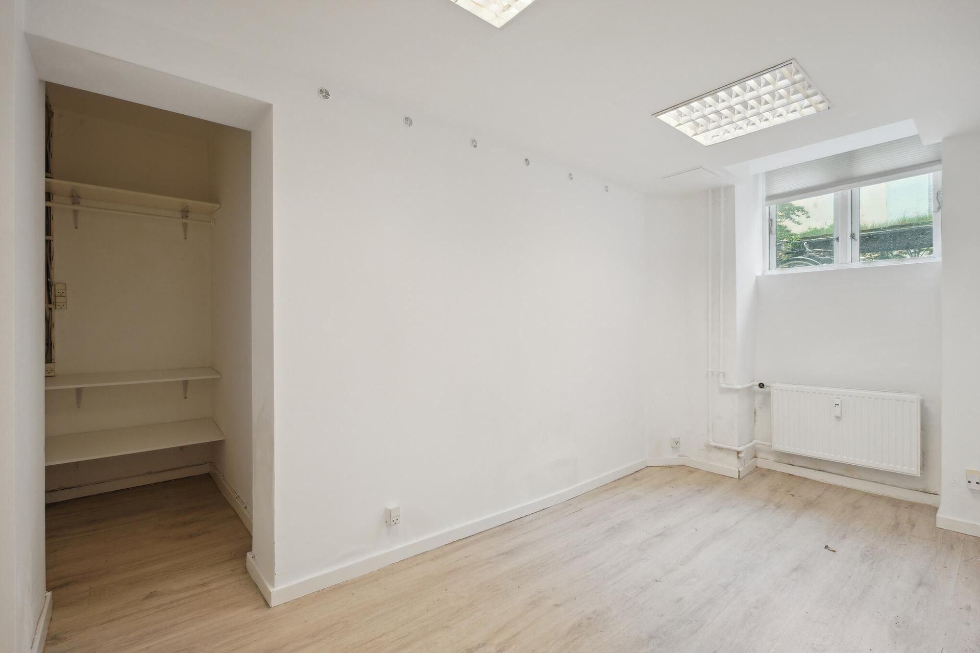 Ledig kælderlejemål med hvidt loft og gulvtæppe, slank garderobe og radiatorer på væggen. Lyst og nytteløst rum til kontor