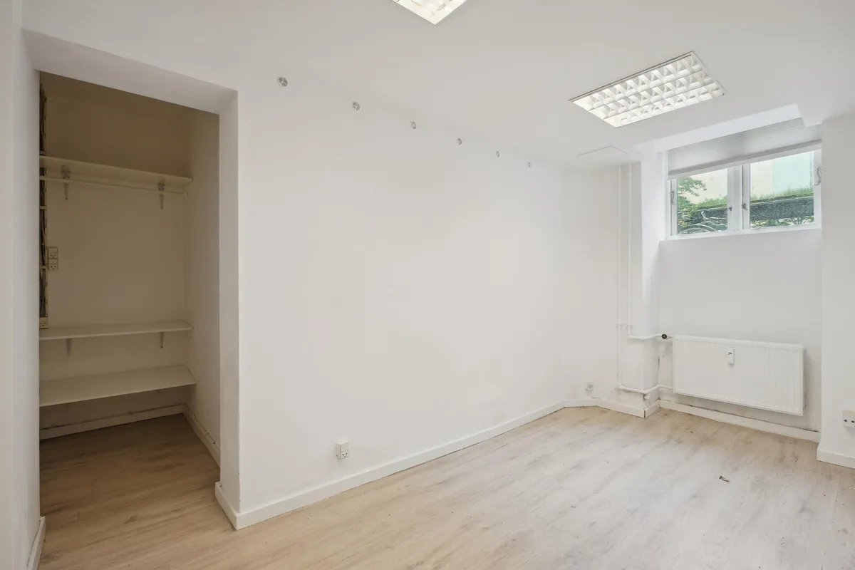 Ledig kælderlejemål med hvidt loft og gulvtæppe, slank garderobe og radiatorer på væggen. Lyst og nytteløst rum til kontor
