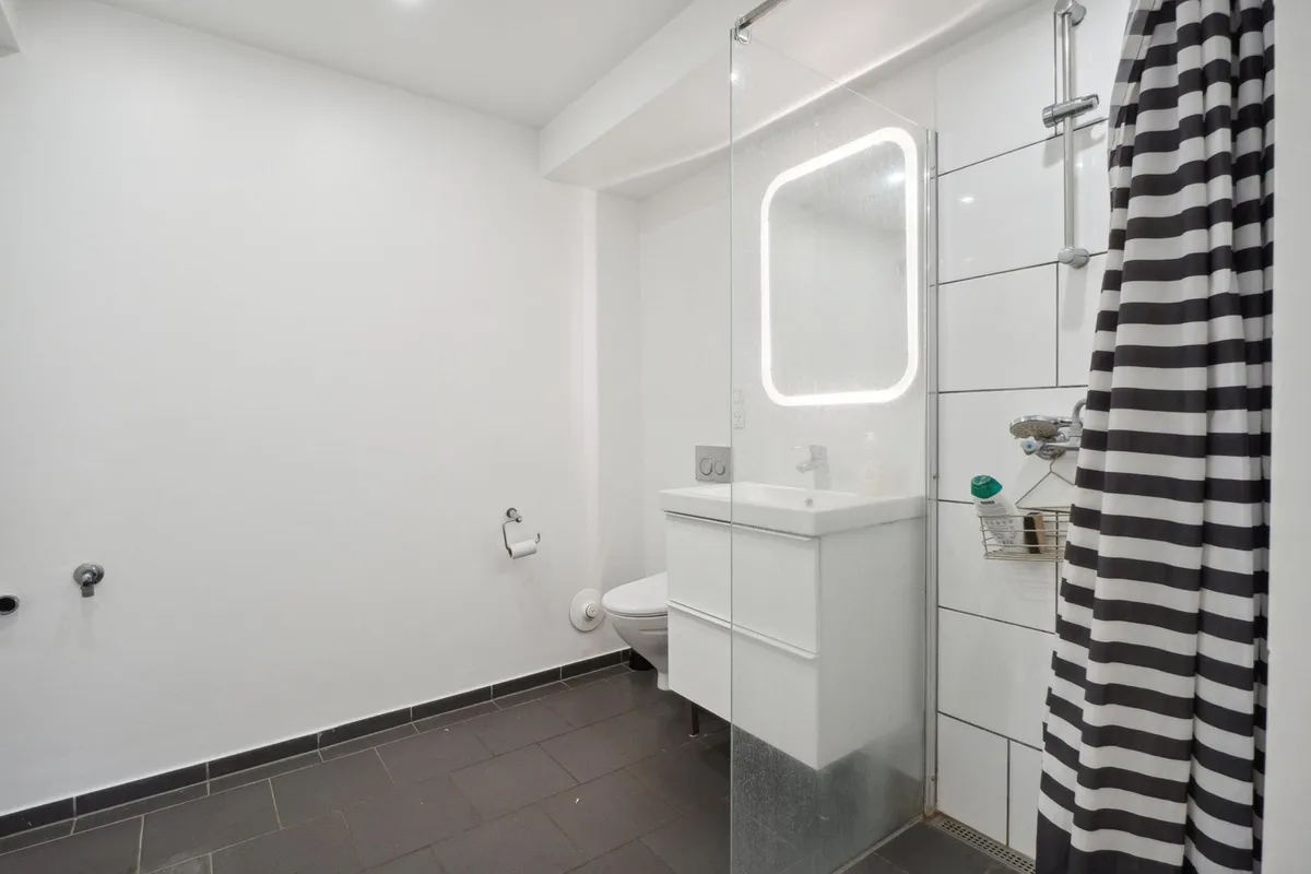 Lejlighed med lyse hvide vægge og gulv og en hvid simpel badeværelse med brusekabine og spejl