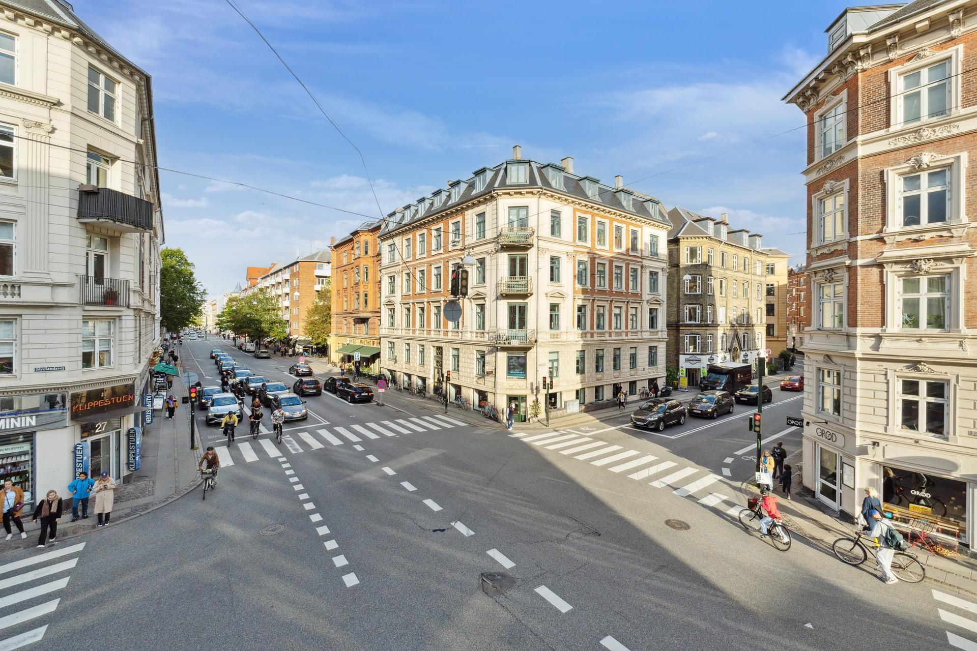 Gadekorsning med folk og cyklister, slanke bygninger med mange vinduer, røde biler og en bus, vejsiderne med forretninger og