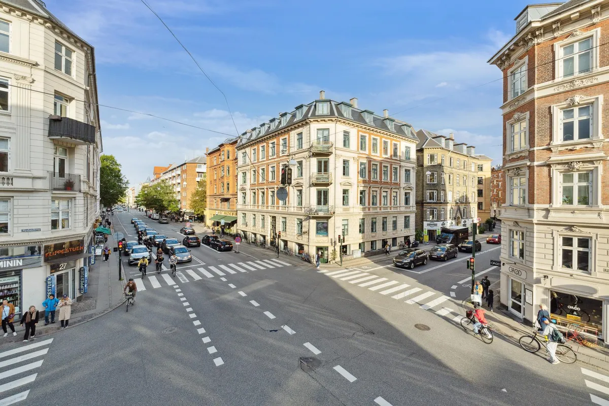 Gadekorsning med folk og cyklister, slanke bygninger med mange vinduer, røde biler og en bus, vejsiderne med forretninger og