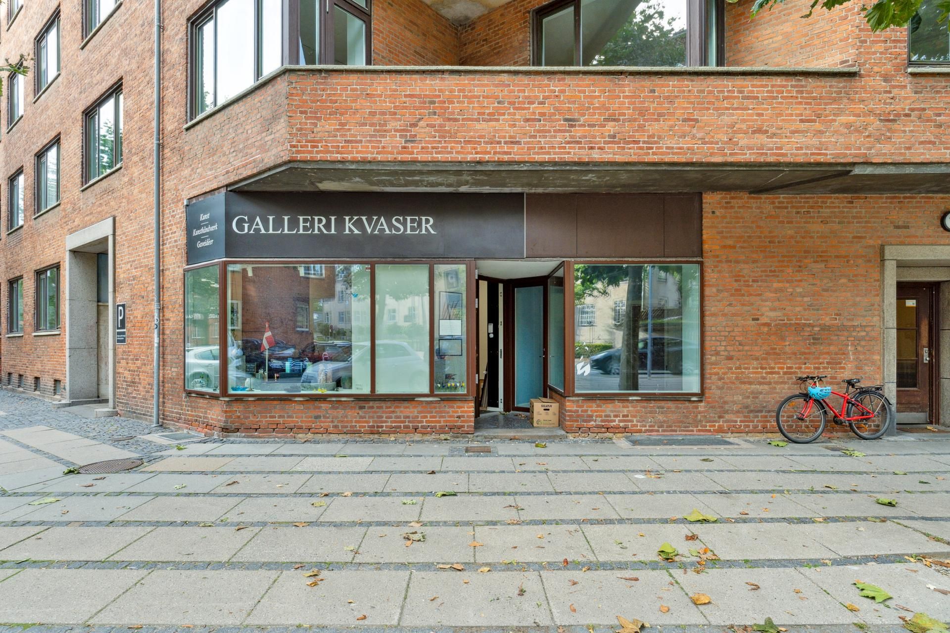Galleri Kvaser på Sankt Nikolaj Vej er indrettet i en rød murstensbygning med flere vinduer og en åben dør. En rødt stel er
