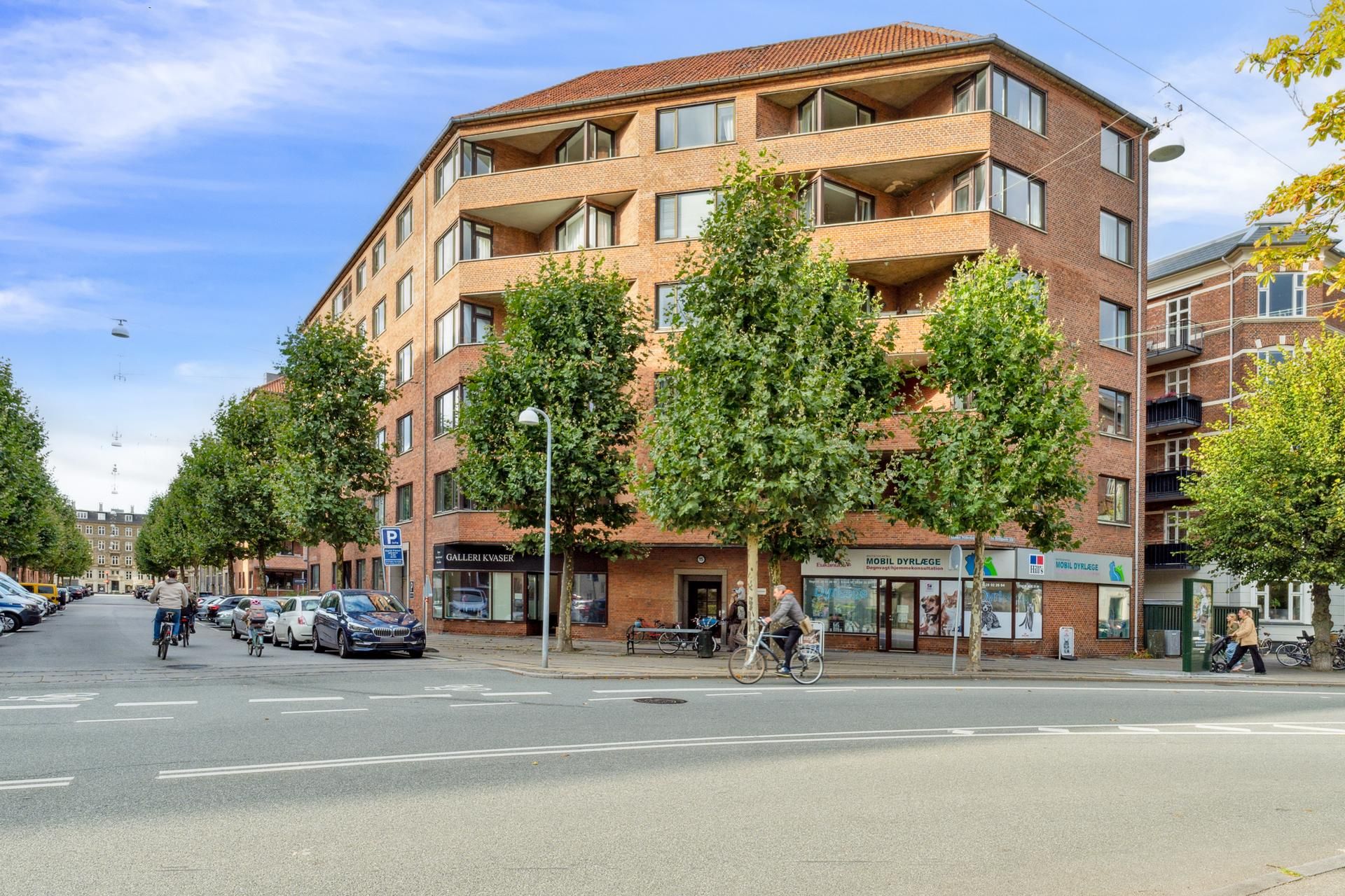 Kvarter med boligbebyggelse og parkeringsplads, to cyklister kører gennem gaden og nogle fodgængere går langs fortovet