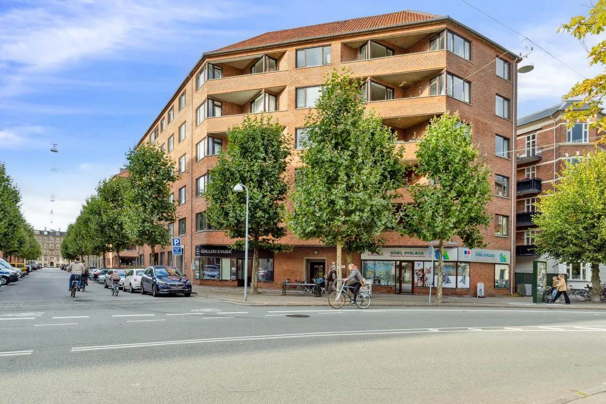 Kvarter med boligbebyggelse og parkeringsplads, to cyklister kører gennem gaden og nogle fodgængere går langs fortovet