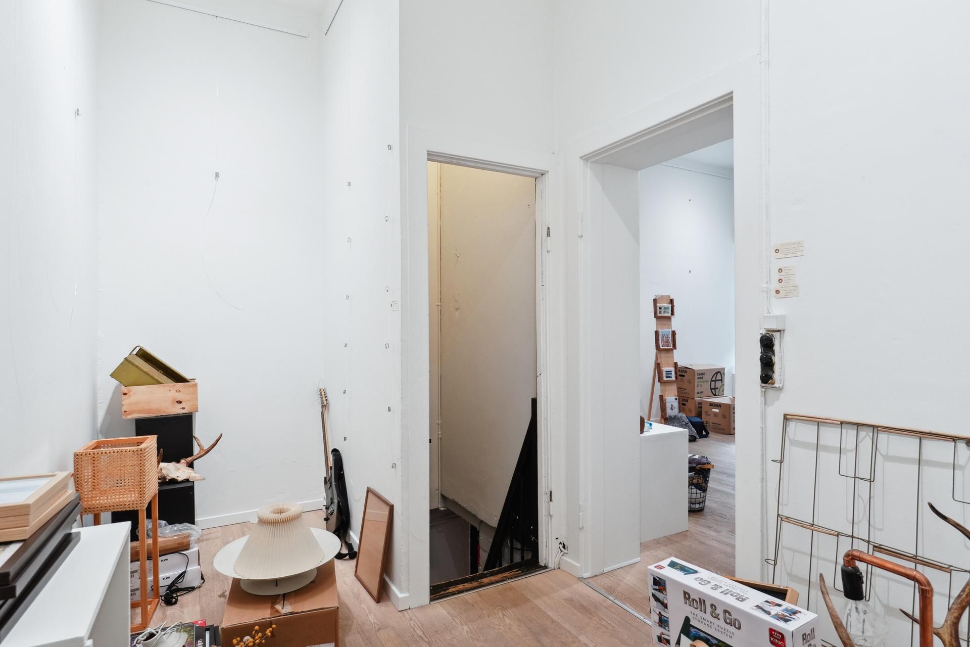 Foto af møbleret lejlighed med forskellige genstande og bokse samt et lukket trappekammer og 2 døre i baggrunden