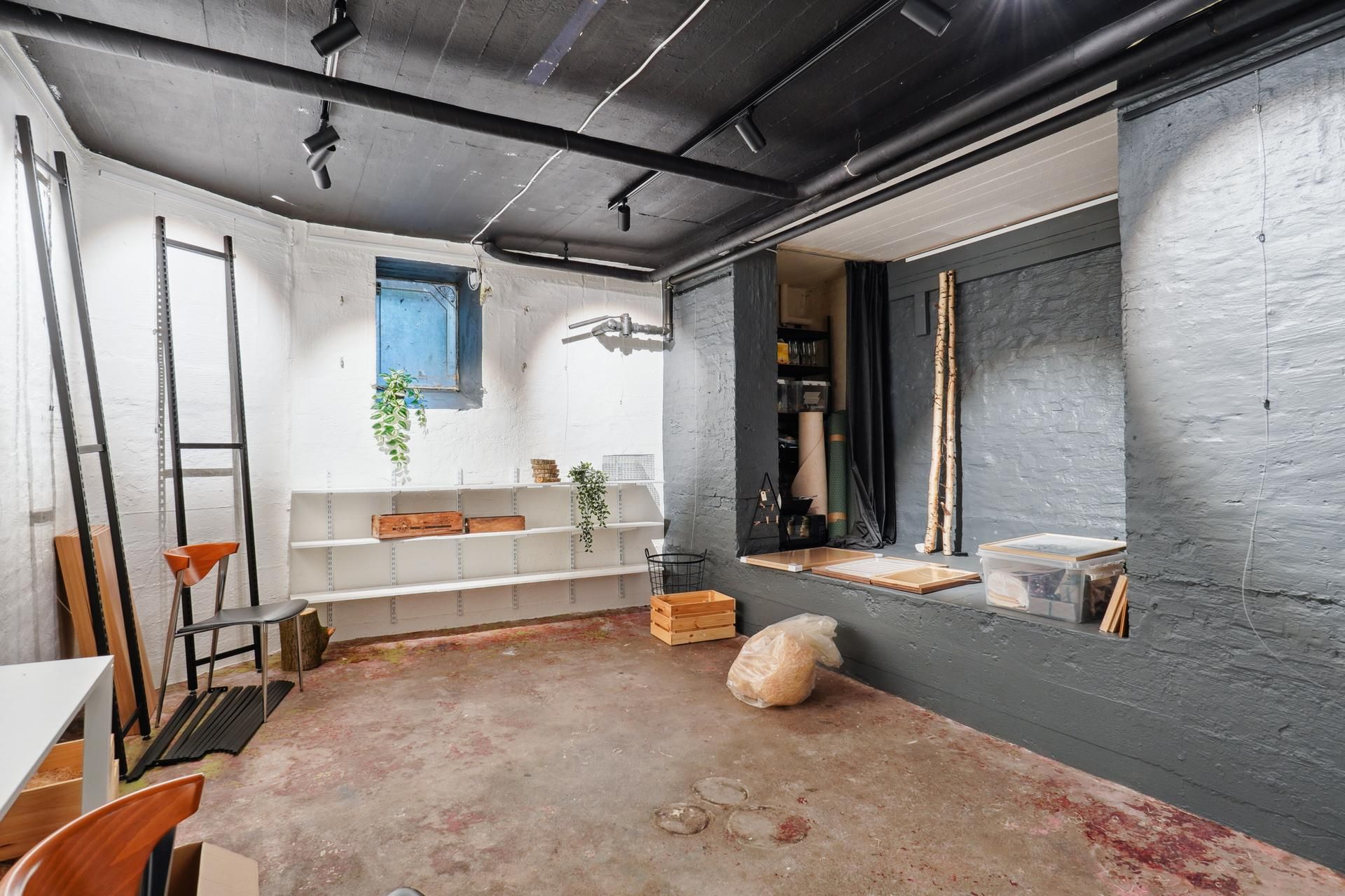 Kælderværelse med højloft, hvide vægge, loftsbjælker og et vindue, der vender ud mod Sankt Nikolaj Vej i København