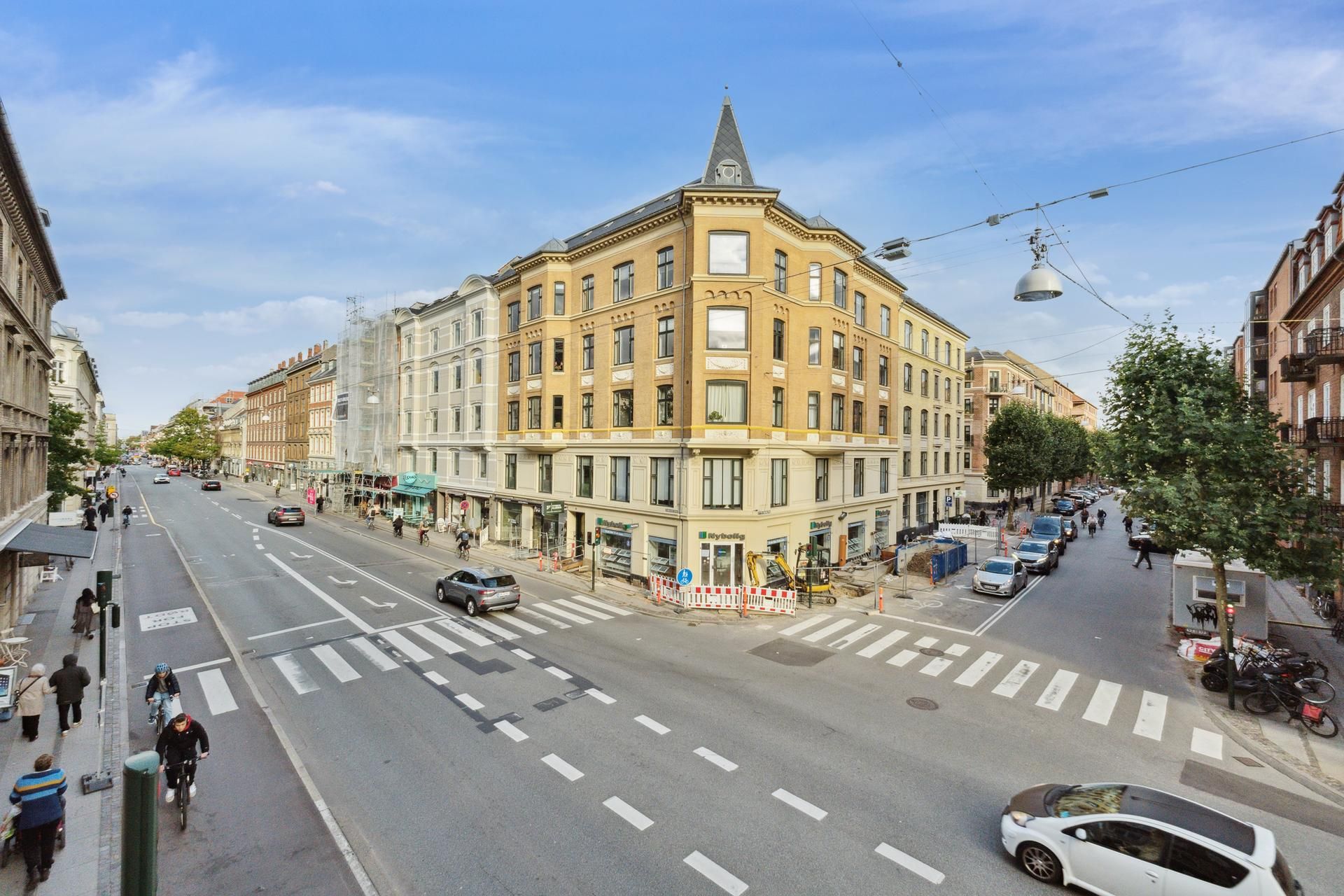 Flere biler og cyklister på Sankt Nikolaj Vej i København med bygninger og grønne træer på begge sider af vejen