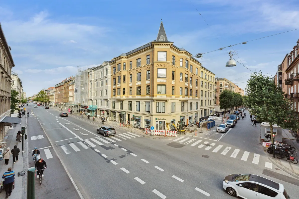 Flere biler og cyklister på Sankt Nikolaj Vej i København med bygninger og grønne træer på begge sider af vejen