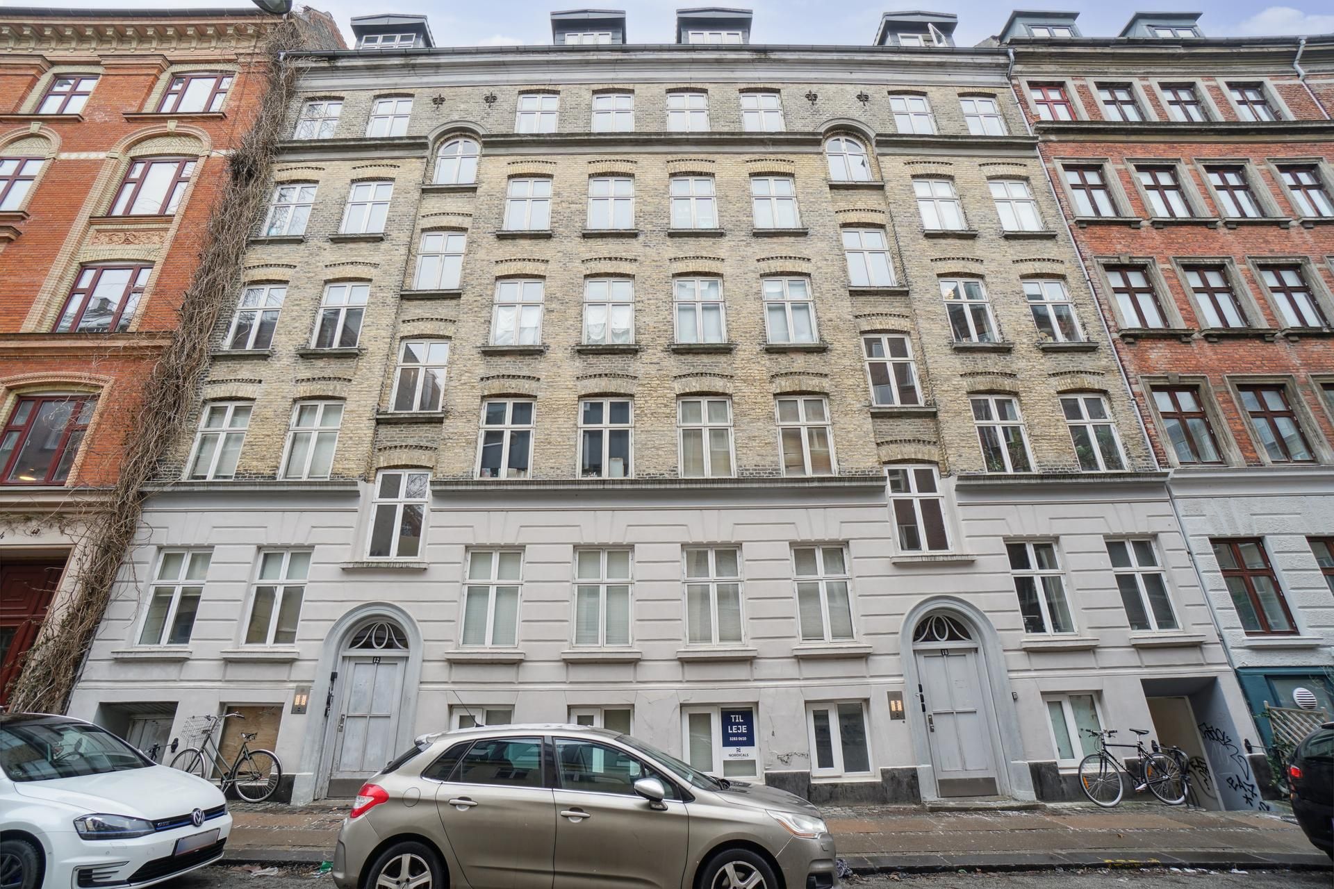 Kælderlejlighed med 120 kvm i gårdmiljø med vinduer, skrånende tag og skarpe linjer. Biler parkeret og cykler ved siden af.