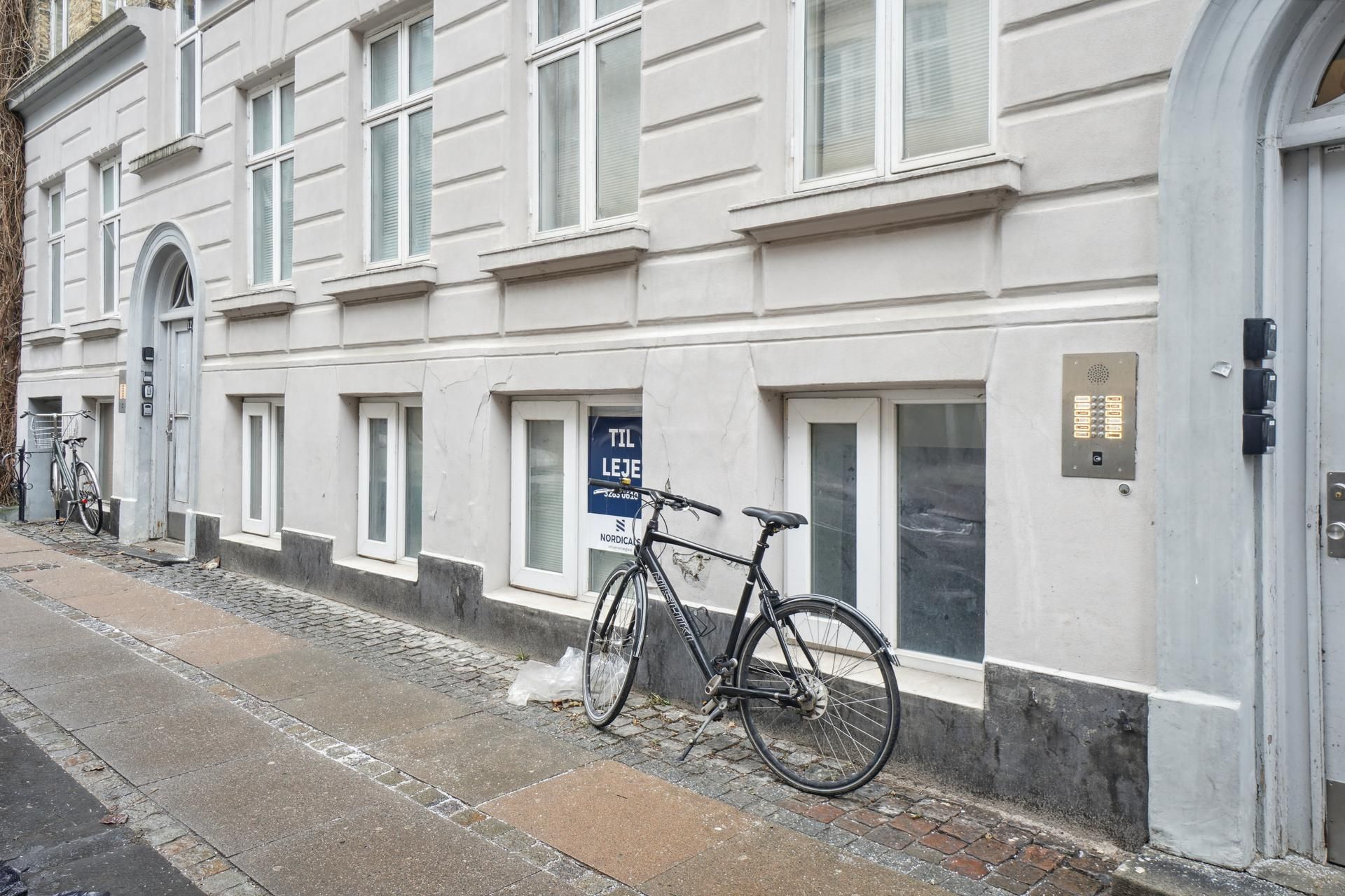 To cykler parkeret udenfor et hvidt bygning med vinduer og en skilt, der læser "Til leje".
