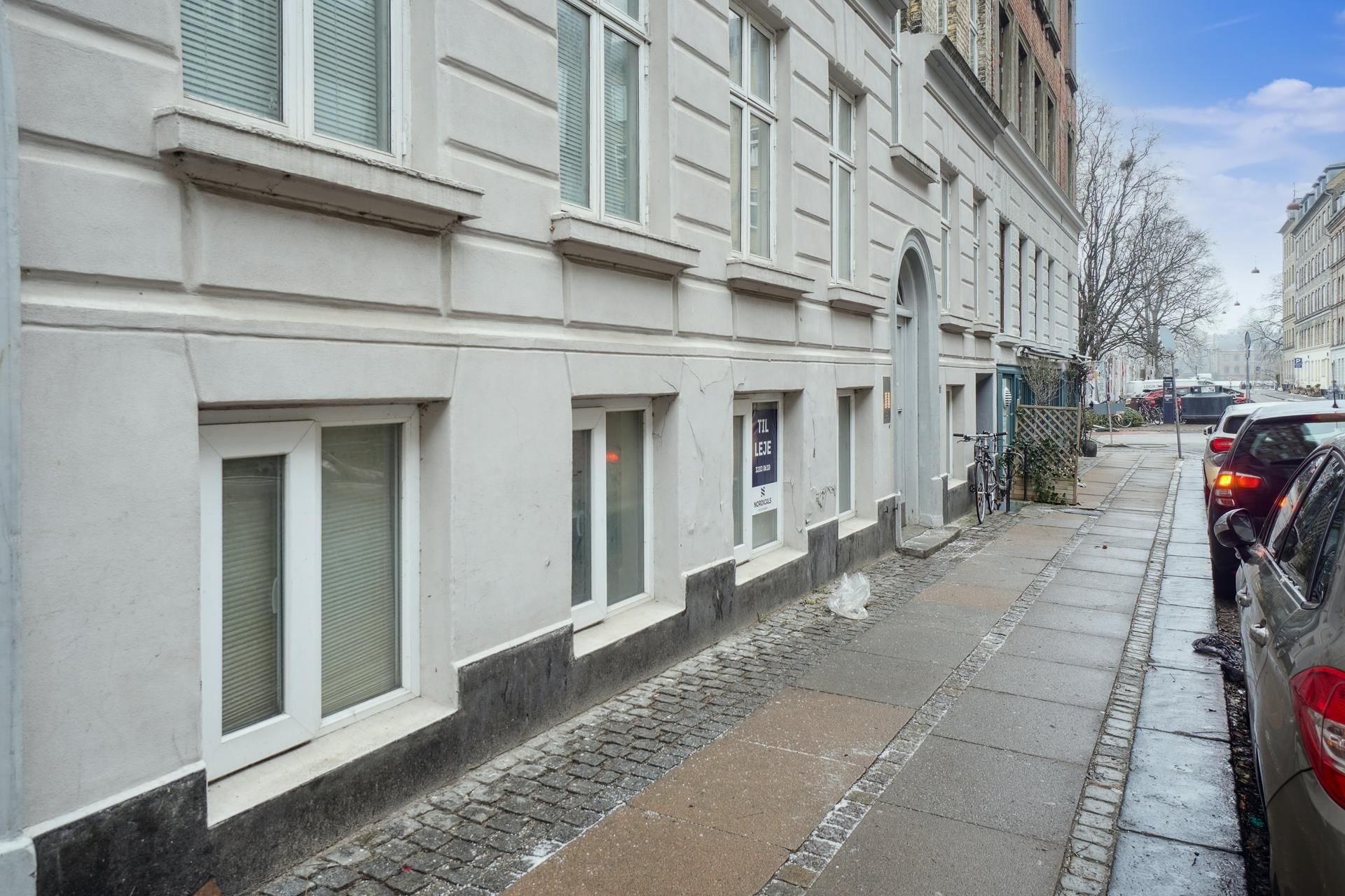 Lejemål med annonceskilt på gadehjørnet i København. Parkerede biler og en cykel langs fortovskanten. Bygningen har hvide