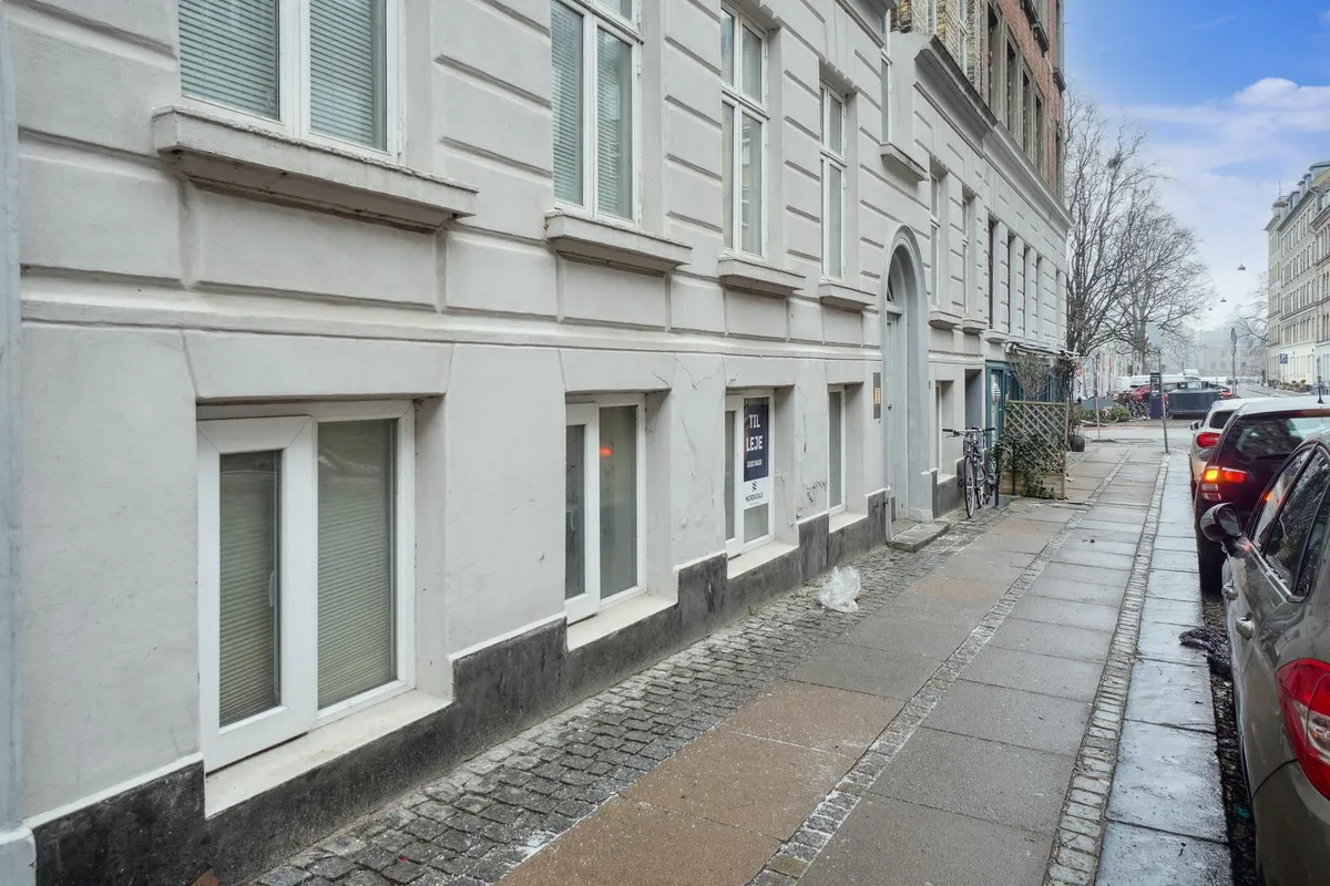 Lejemål med annonceskilt på gadehjørnet i København. Parkerede biler og en cykel langs fortovskanten. Bygningen har hvide