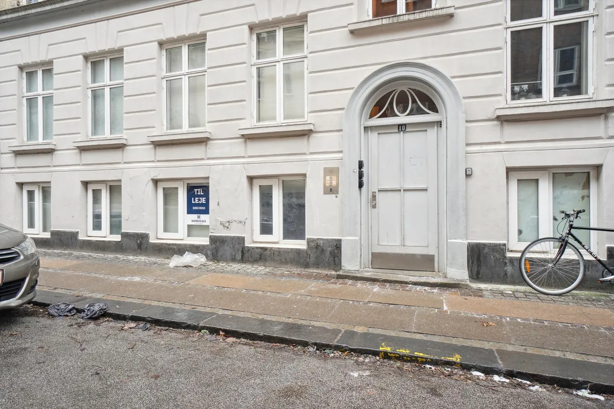 Lejlighed med vinduer og indgangsdør, hvor den ene lejlighed er mærket med "TIL LEJE" og nummeret 10. Cykel og bil parkeret