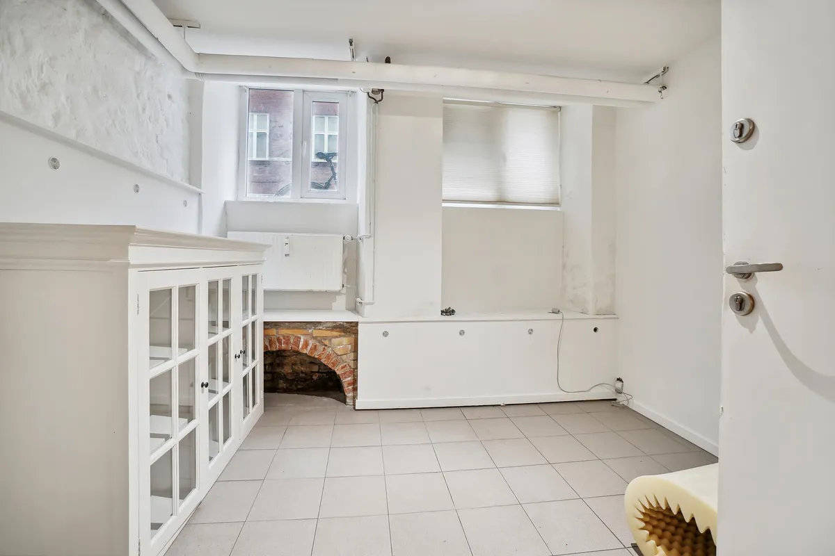 Kælderlejlighed med højloftede rum og hvide vægge, to vinduer, skraldespand og garderobeskab med glasdøre