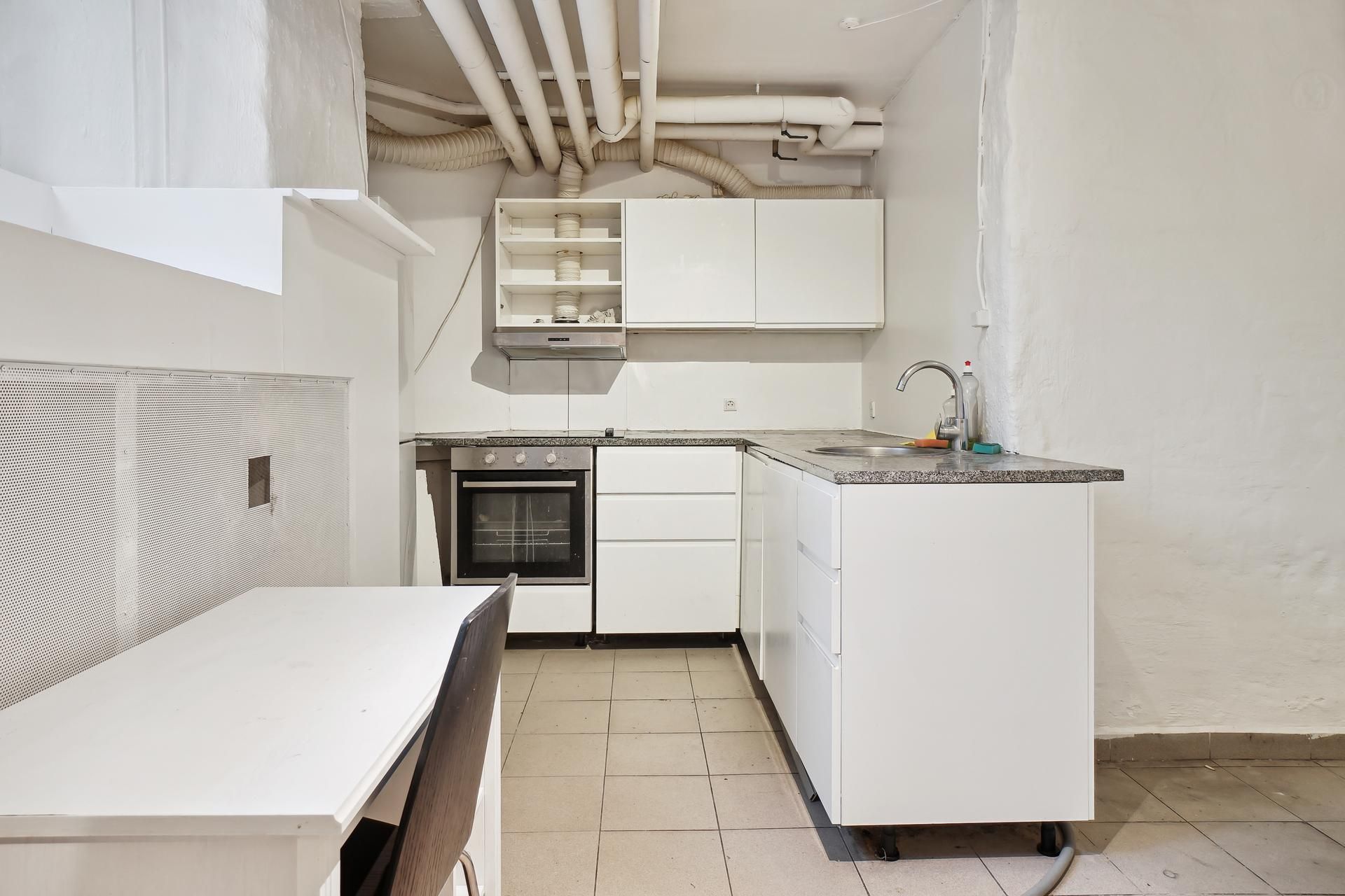 Et minimalistisk køkken med hvide vægge og loft, højt til loftet, hvide skabsfronter, grå gulvfliser, en indbygget komfur,