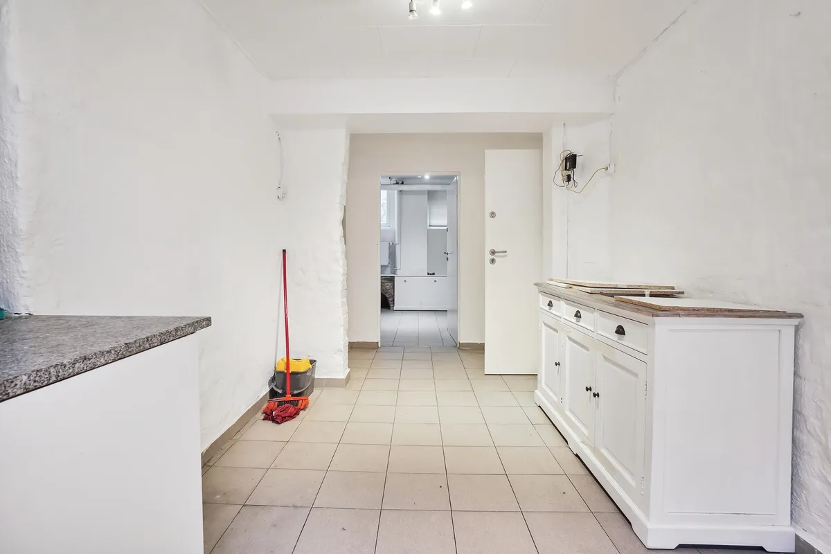 Indvendigt i et kælderlejemål er der lyse lofter, hvidt køkken og tømredarbejde. Et håndvask i køkkenet og en vaskemaskine.