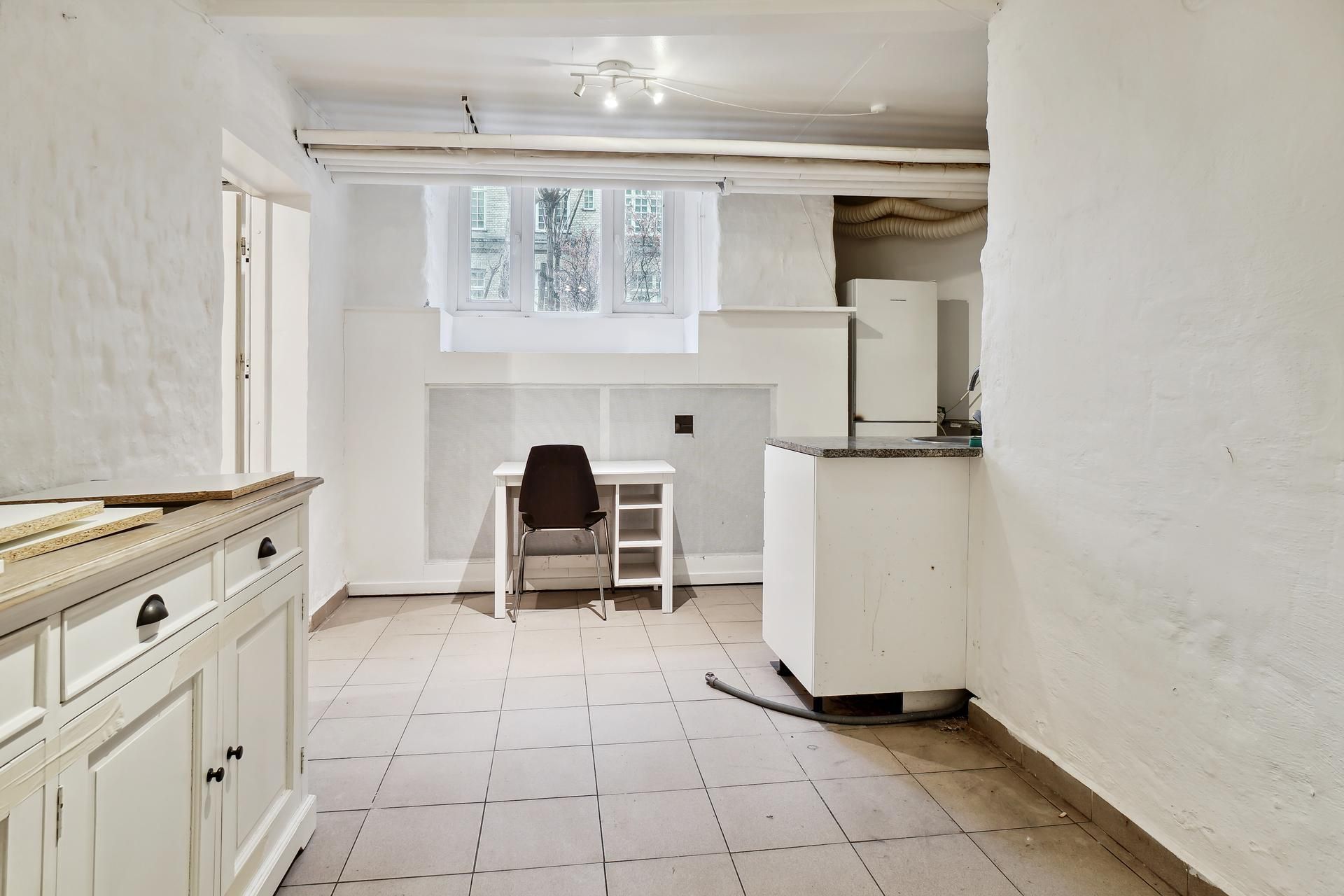 Kælderlejlighed med lyse loftsvinduer, lysekroner og lysegrå lofter, lysegule vægge, hvidt køkken, hvidt bord og stol samt