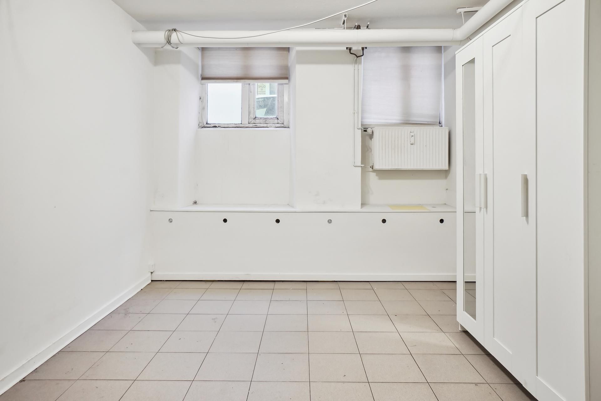 Tomt kælderlejemål med højt til loftet, to vinduer og et hvidt vindue, kabinetter og radiatorer.