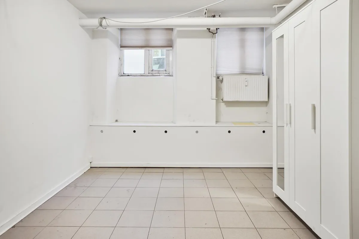 Tomt kælderlejemål med højt til loftet, to vinduer og et hvidt vindue, kabinetter og radiatorer.