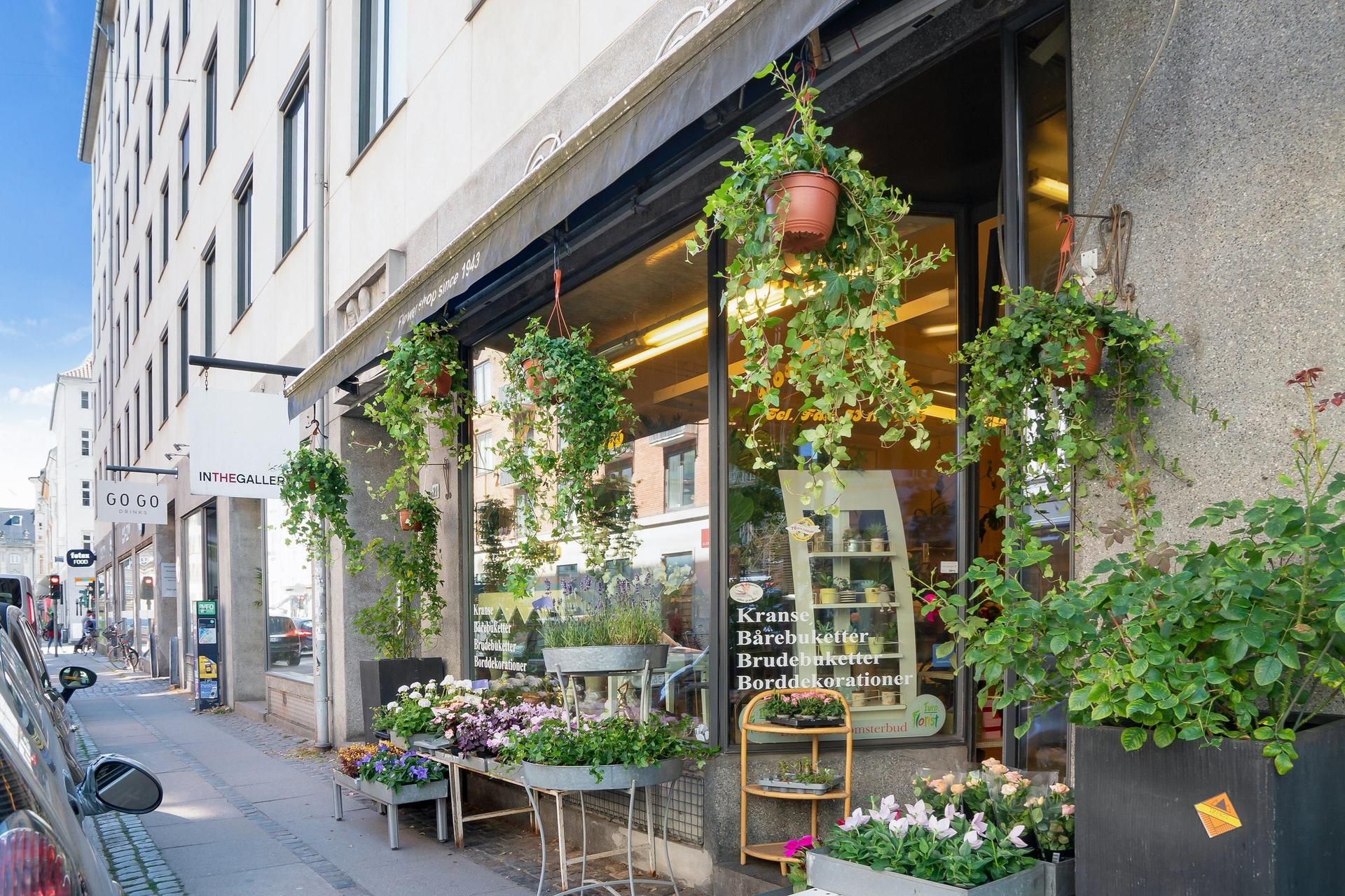 En butik med planter og blomster i potteplanter, udendørs. Sort banner med teksten Kranse, Bærebøjetter, Brudebøjetter og