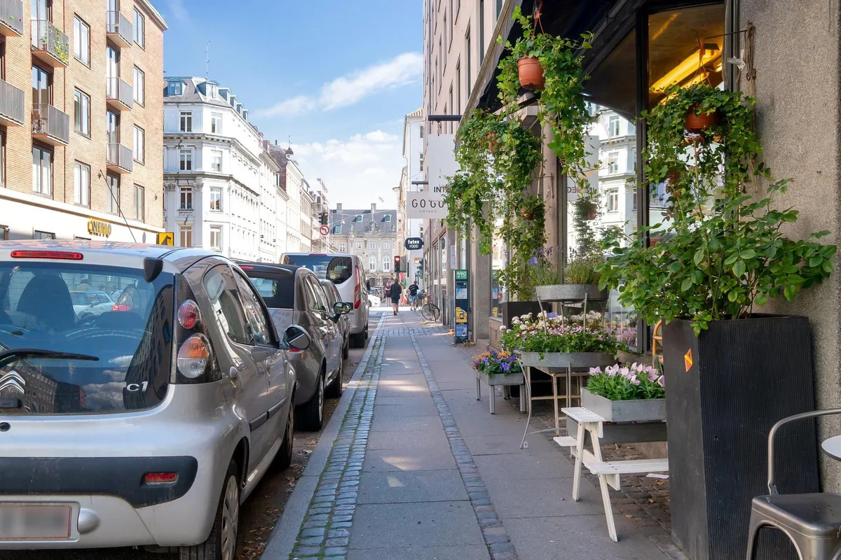 En bred gågade med parkerede biler og blomsterpotter samt en restaurant med træstole og terrassemøbler