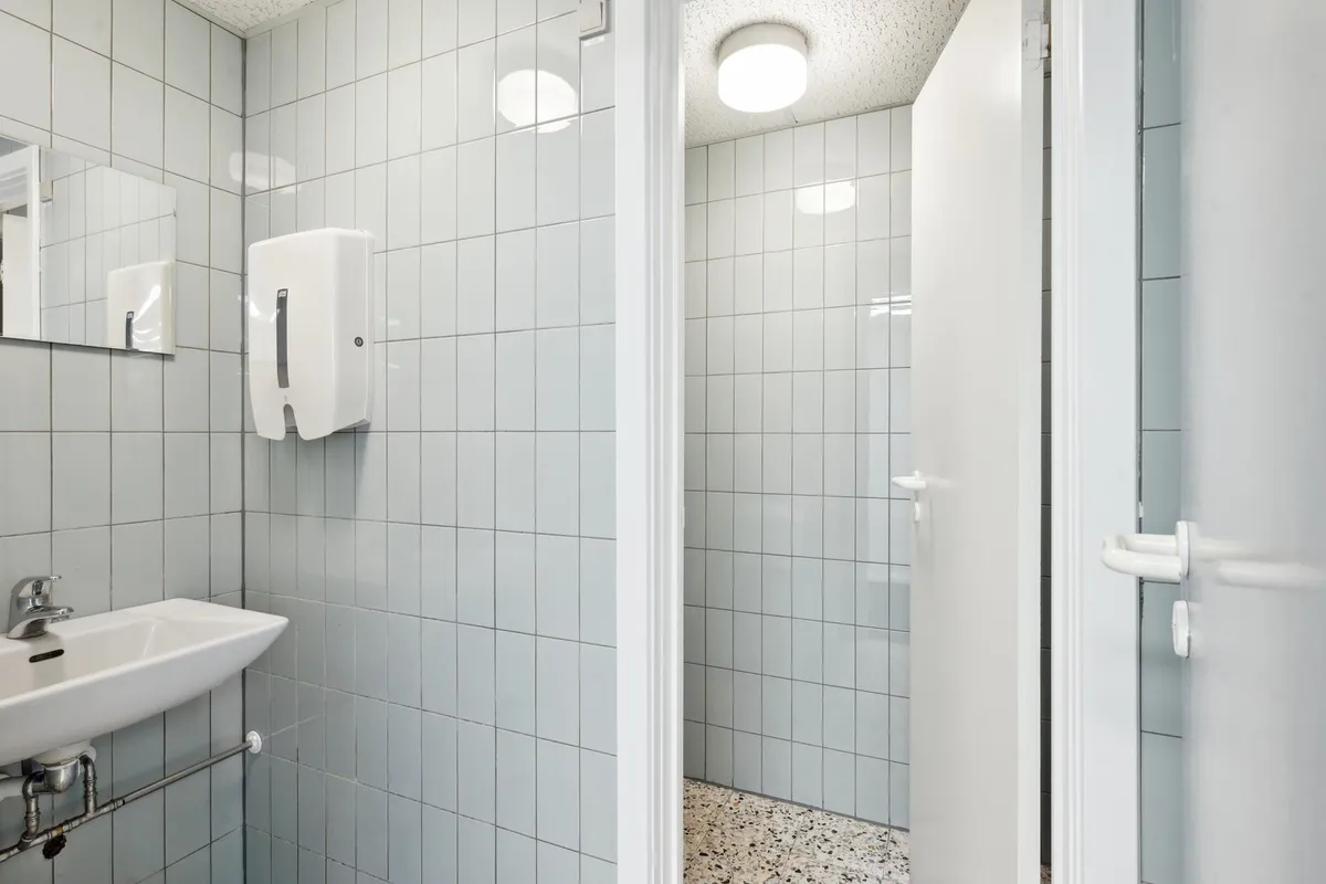 Et rent og enkelt badeværelse med hvidt flisegulv, en hvid vask, en håndklædeautomat og et lysekronlys på loftet