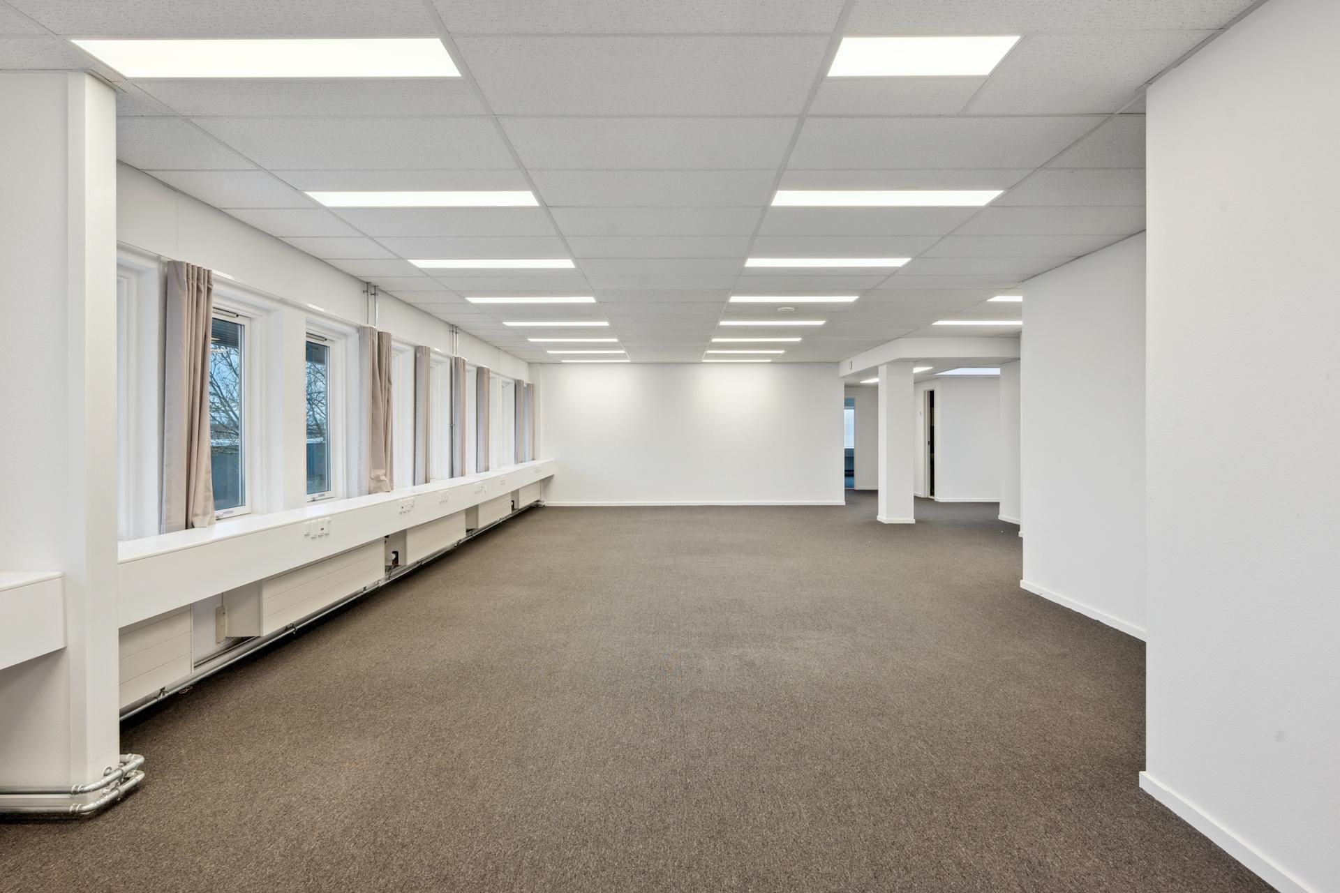 Kontor med hvide vægge, lysegrå gulvbelægning og højloft med indbygget lys. Værelset har lysegrå gardiner og et hvidt panel