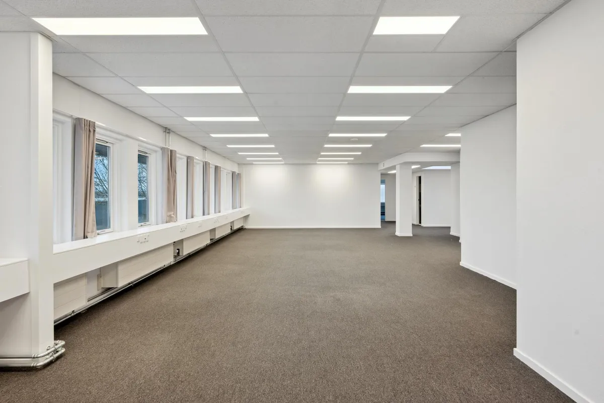 Kontor med hvide vægge, lysegrå gulvbelægning og højloft med indbygget lys. Værelset har lysegrå gardiner og et hvidt panel