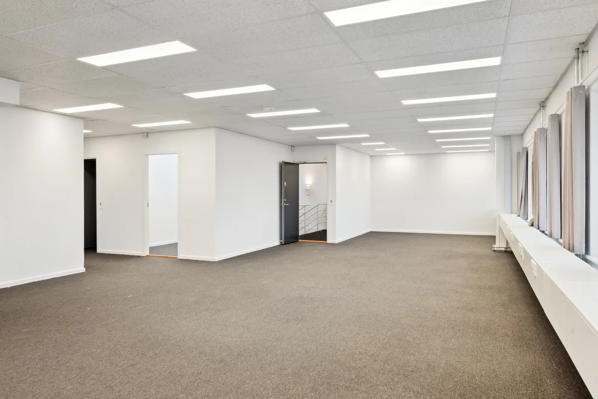 Rummelig og lys kontorlokal med lysegulv og hvide vægge. Lokaler med flere indgange, hvoraf én er åben. Værelserne er tømt