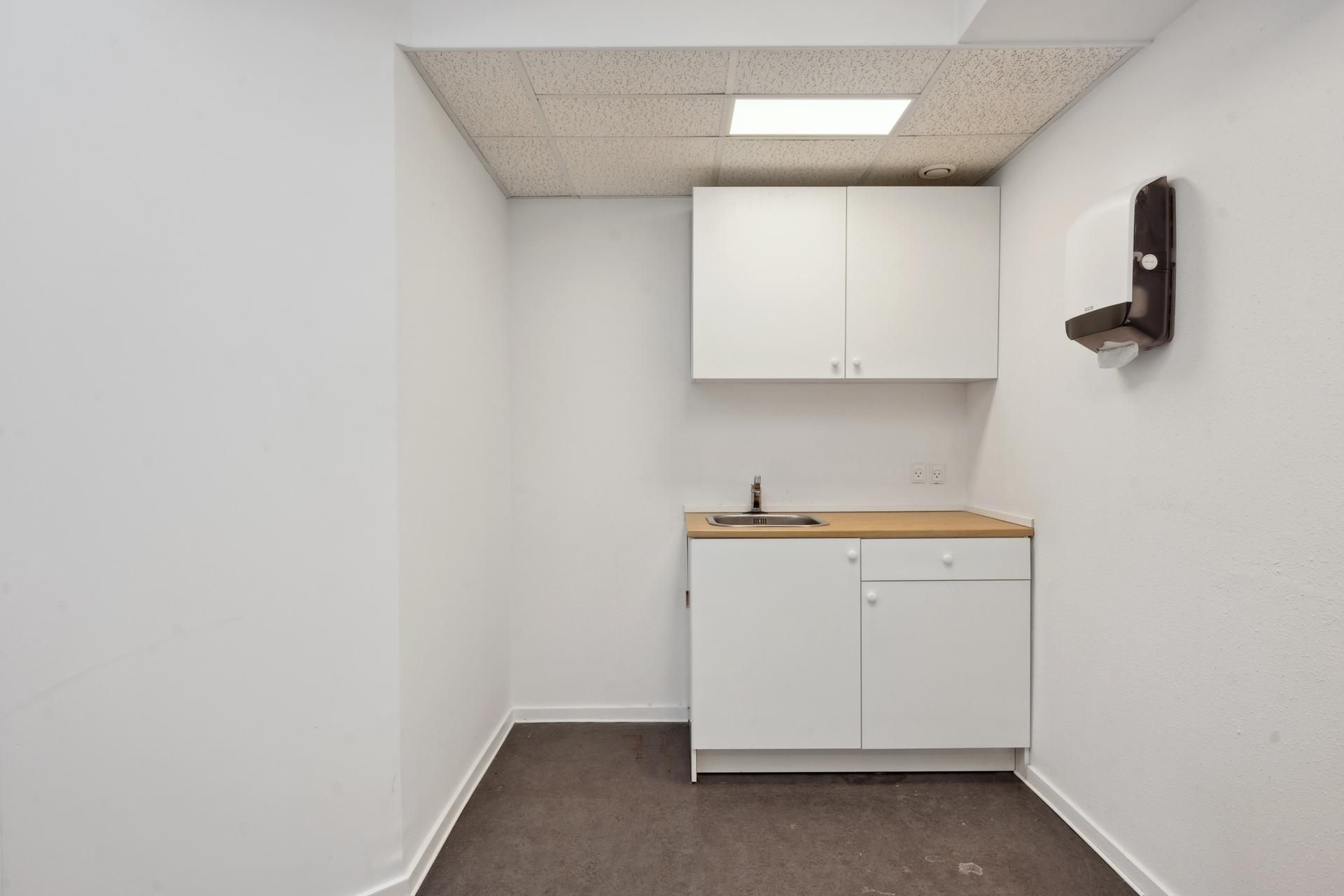 En tom, hvidmalet kontorværelse med trægulv og en hængende håndklædeautomat samt et højreguleret højskab med vaskebækken og