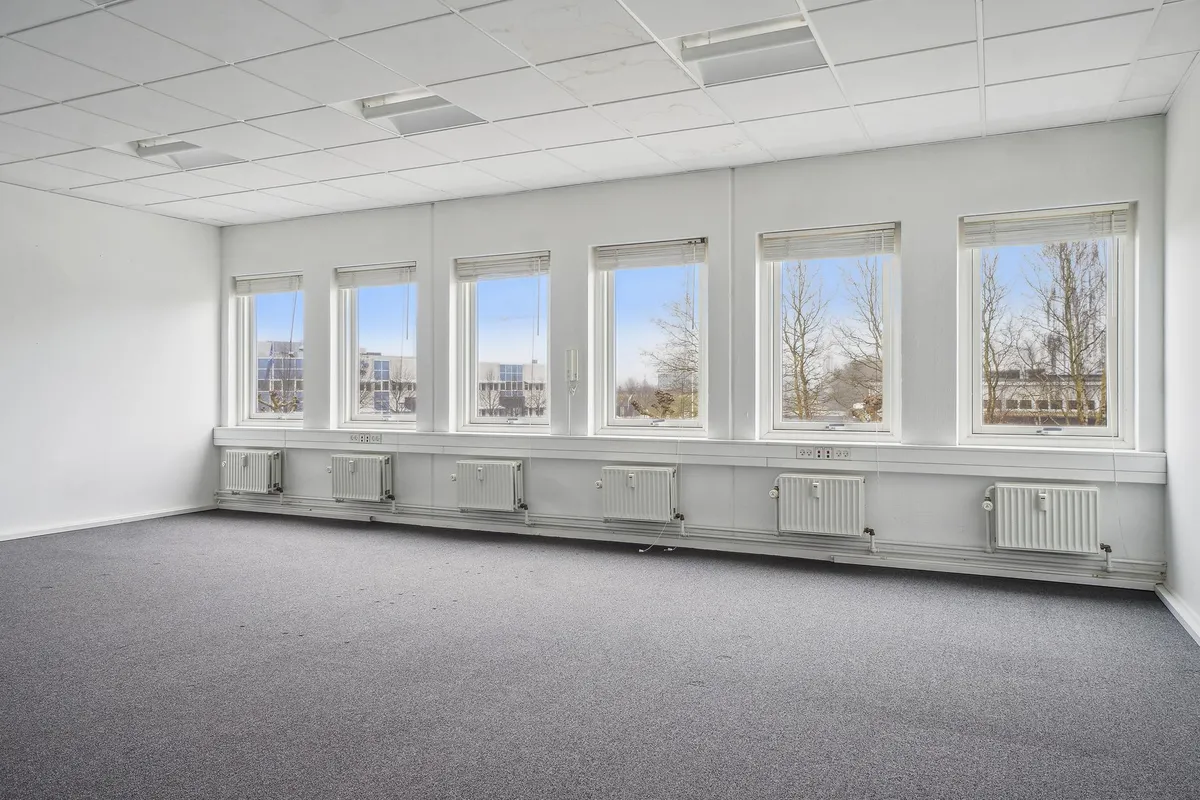 Et stort, lyst og velholdt kontorlokale med hvide vægge, grå gulvbelægning og vinduer med gardiner.