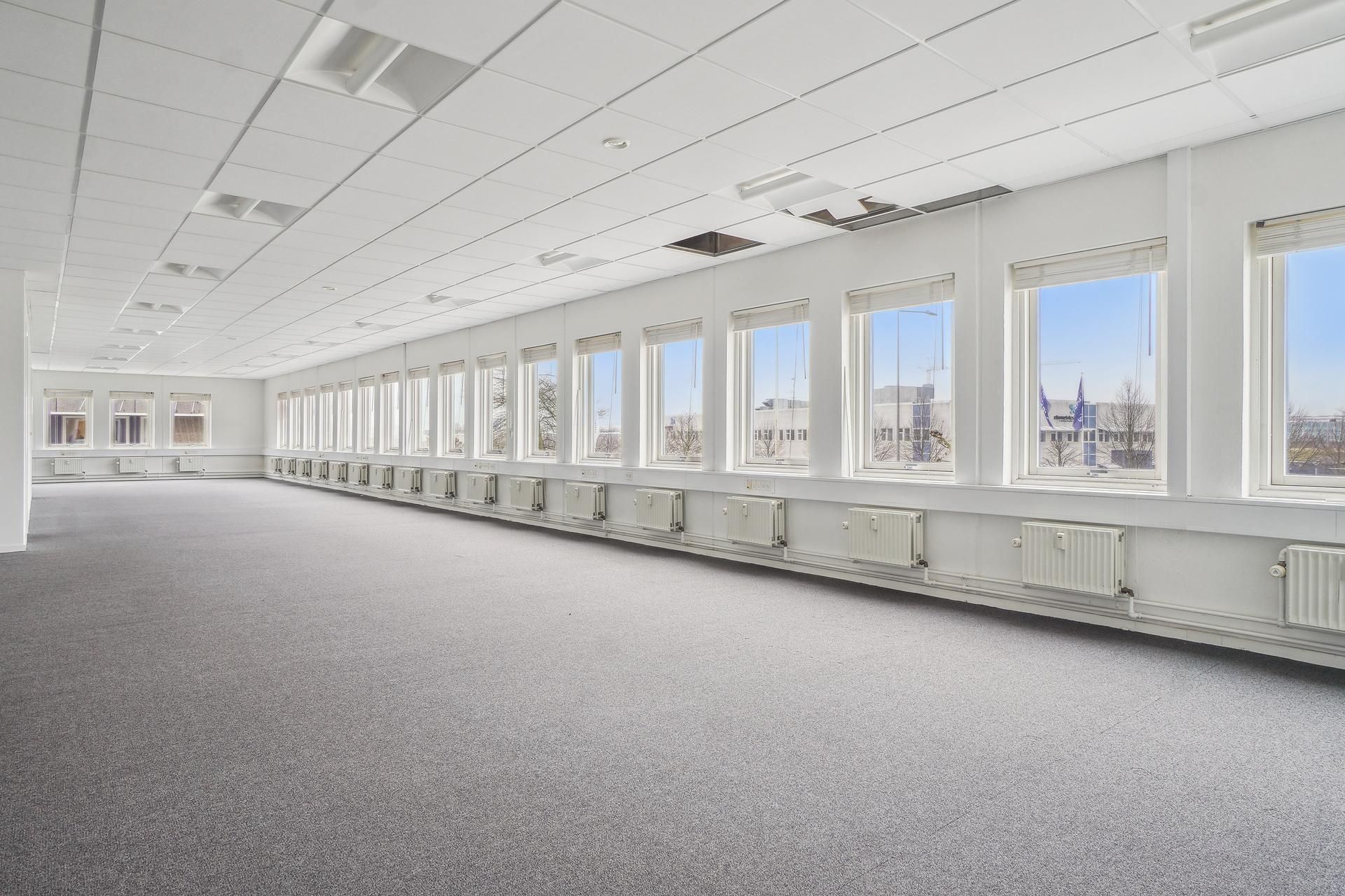 Lyst og velholdt kontorlokal med store vinduer og hvide vægge samt højt til loftet og lyse loftsplader