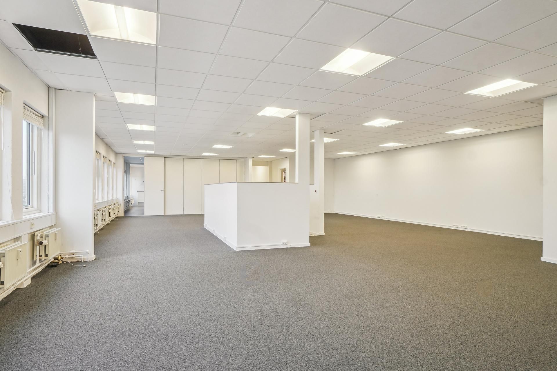 En lys kontor med hvide vægge, gulv og loft, hvor gulvet er dækket af stof og der er store vinduer og hvide døre.