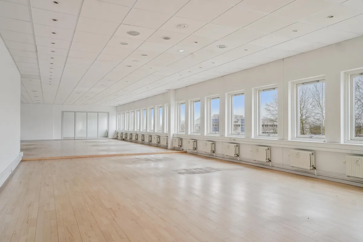 Lyst kontor med lyse gulve, hvide vægge og store vinduer med udsigt til bygninger og træer
