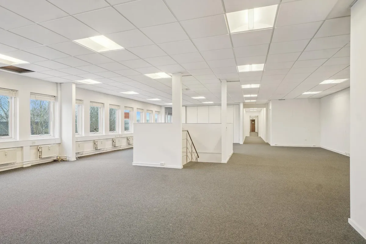 En lys og velholdt kontorlokal med store vinduer og hvide vægge samt lyse loftsbeklædninger og en slank stige