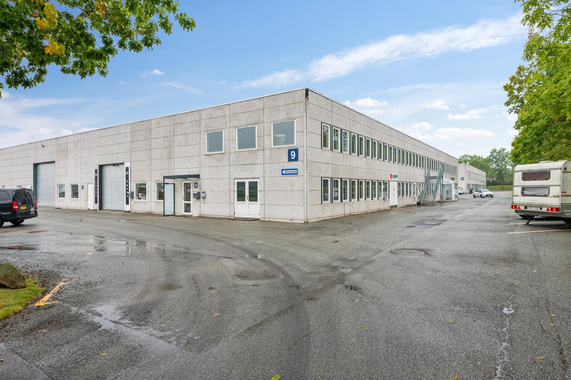 En stor hvid bygning med flere garageindkørsler og vinduer er parkeret med biler og en campingvogn på den ene side og en SUV