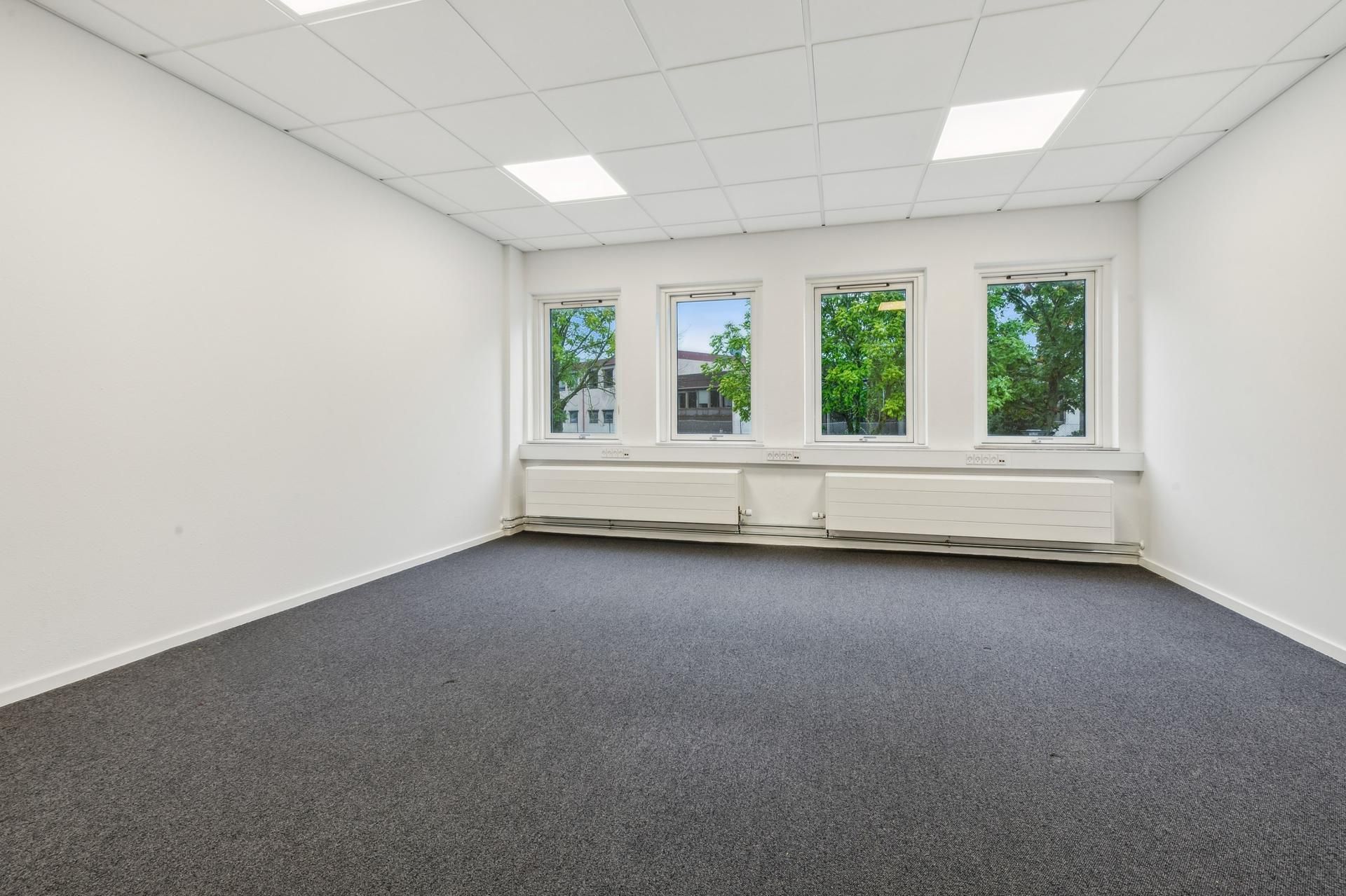 Kontoretage med hvide vægge, gulv og loft, tre vinduer med gardiner og udsigt til grønne træer, radiatorer under vinduet,