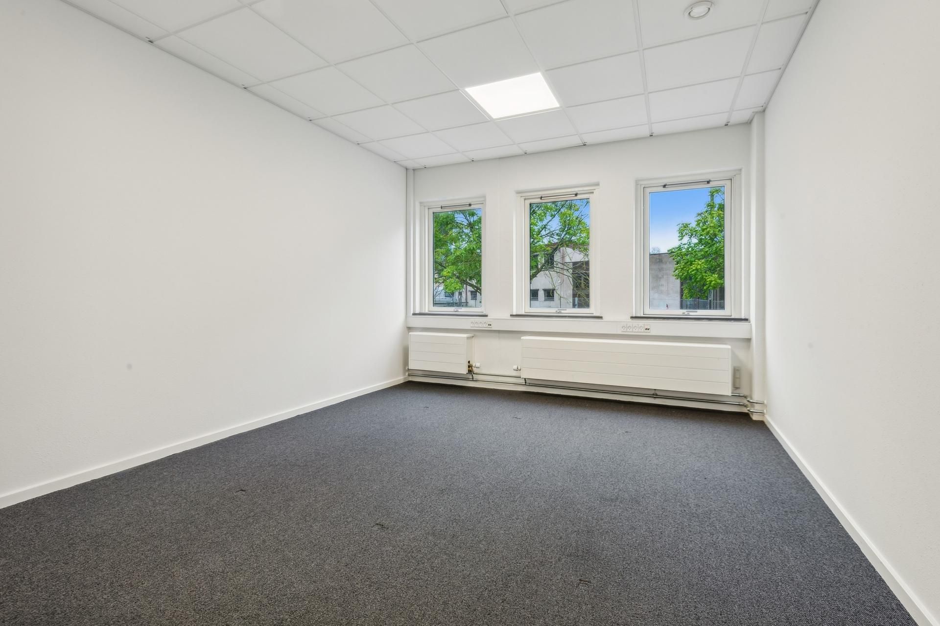En tom og nyrenoveret kontorvirksomhed med hvide vægge, højt til loftet og lyse gulvbelægning. Der er tre store vinduer, der