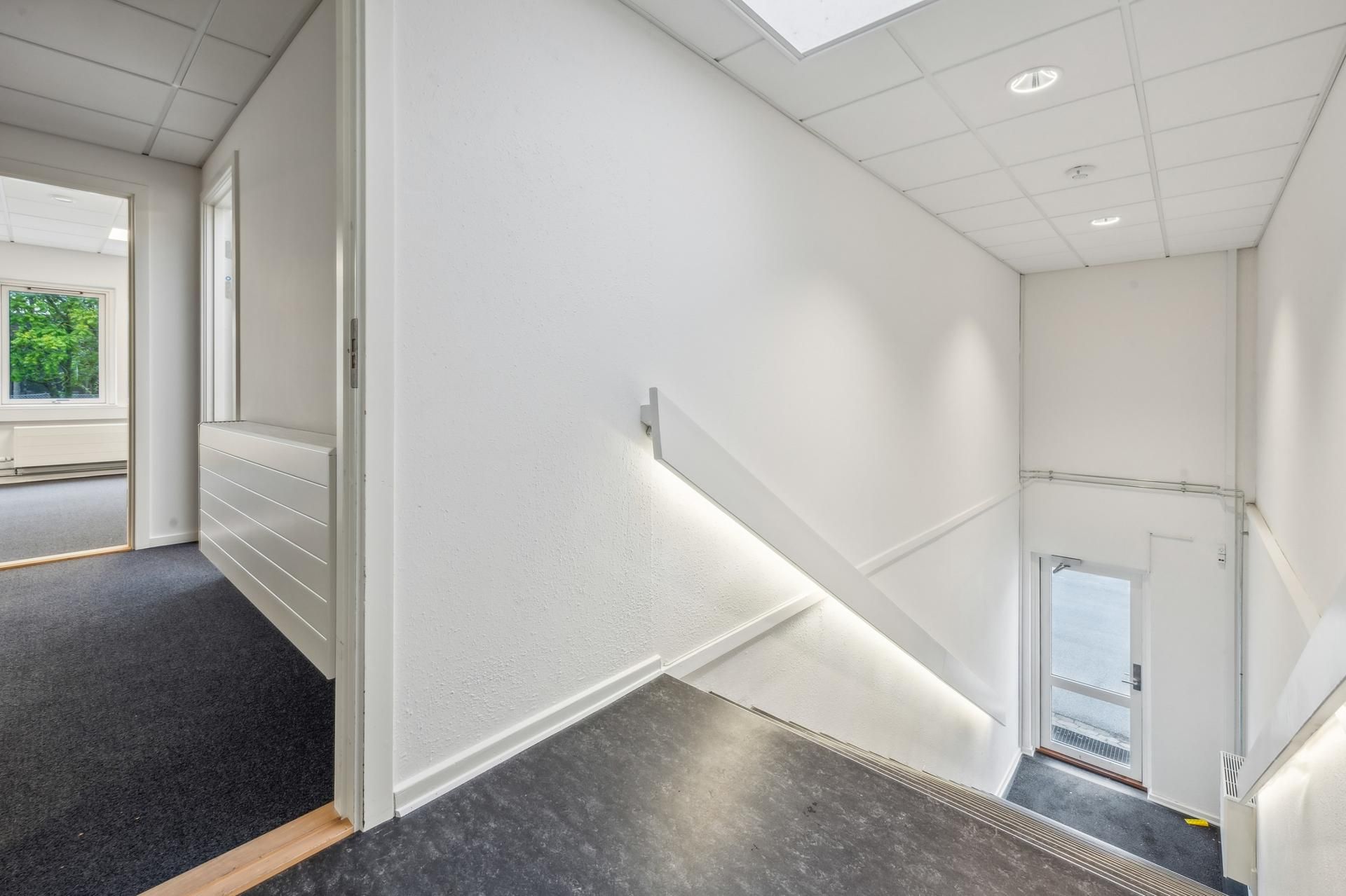 Hvid kontoretage med tomt gulv, åben dør til siden og stige til næste etage. Lyse loftspaneler, lyse og hvide vægge.