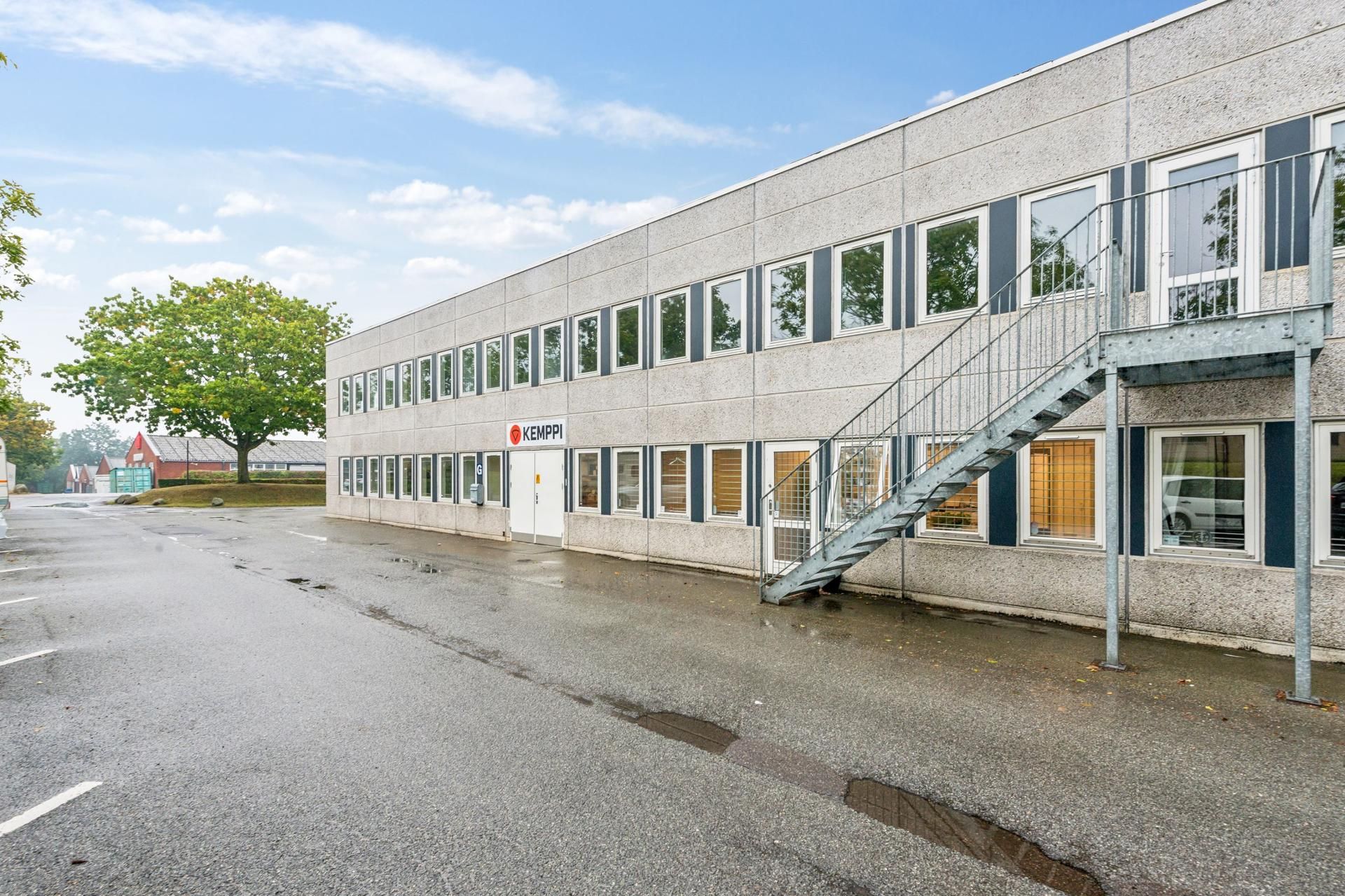 Kontoretage med glasruder, gulv og skrånende trappe til entree. Tegn, der siger Kemppi. Træer og en bygning i baggrunden.