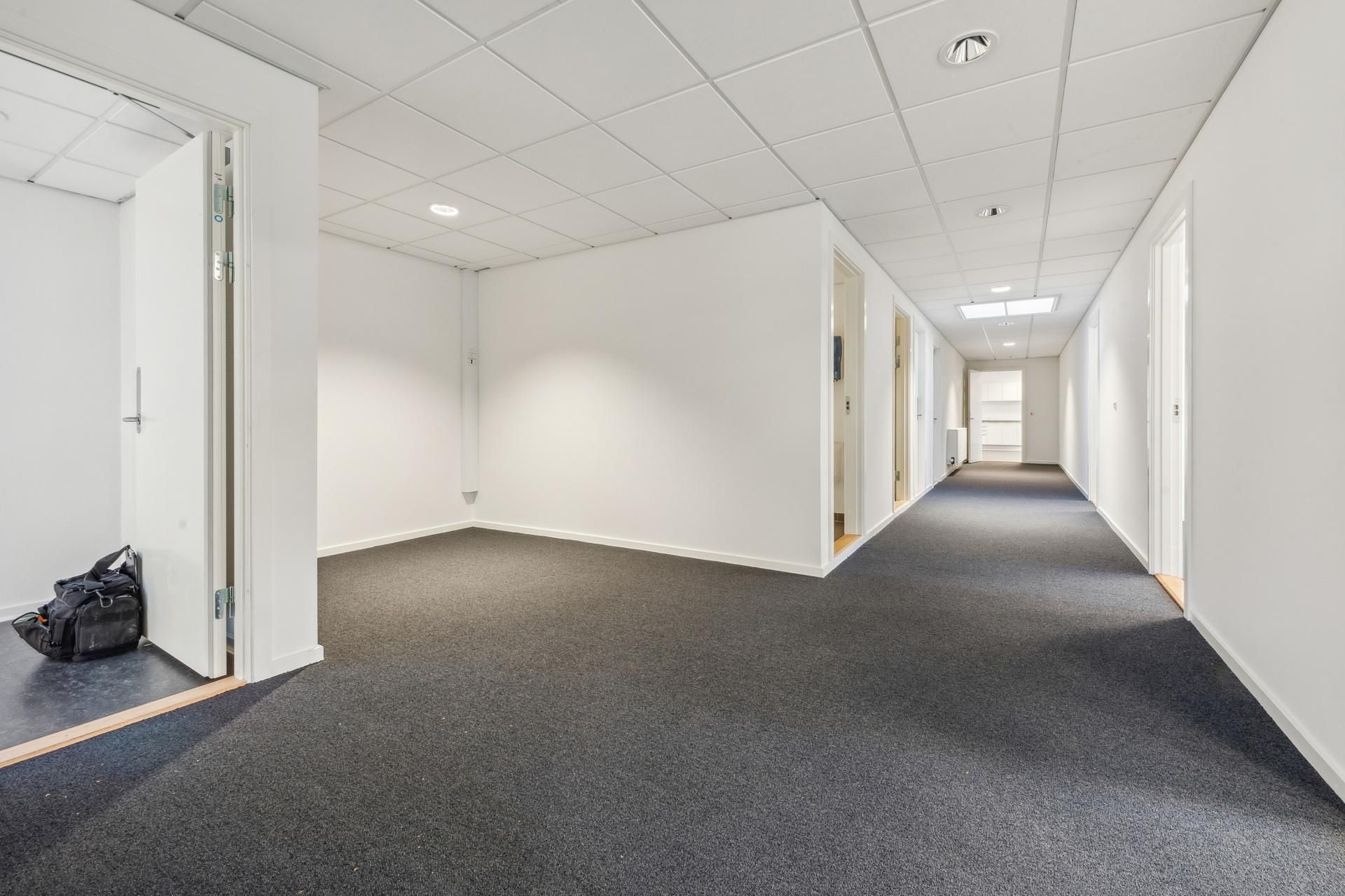 Langt, lyst og rent kontor med hvide vægge og mørke gulve. På højre side er en åben dør, og der er en taske på gulvet til