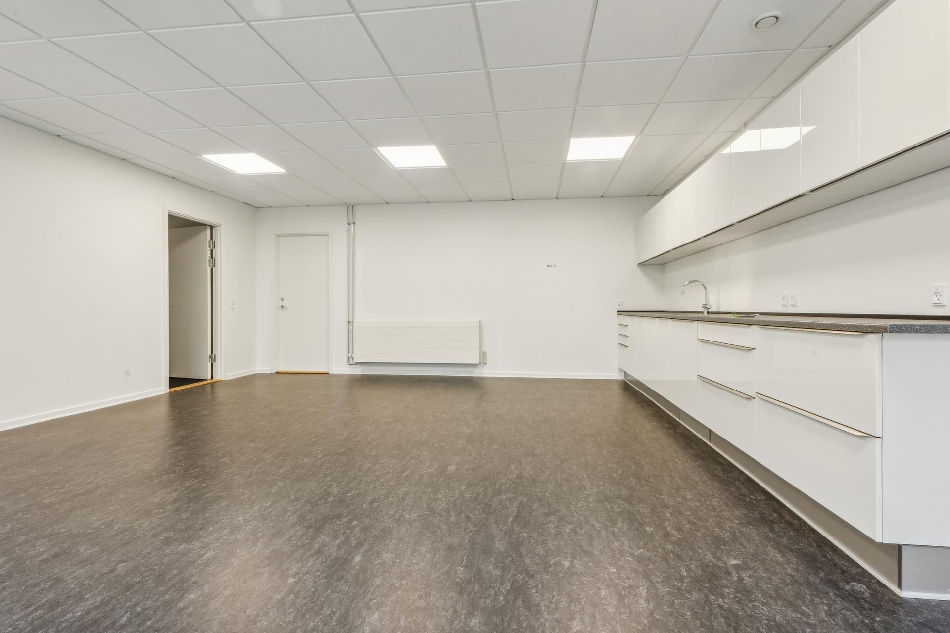 Moderne kontor med hvide vægge, hvide skabe og lysegul gulvbelægning. Der er en dør og et radiator på væggen.